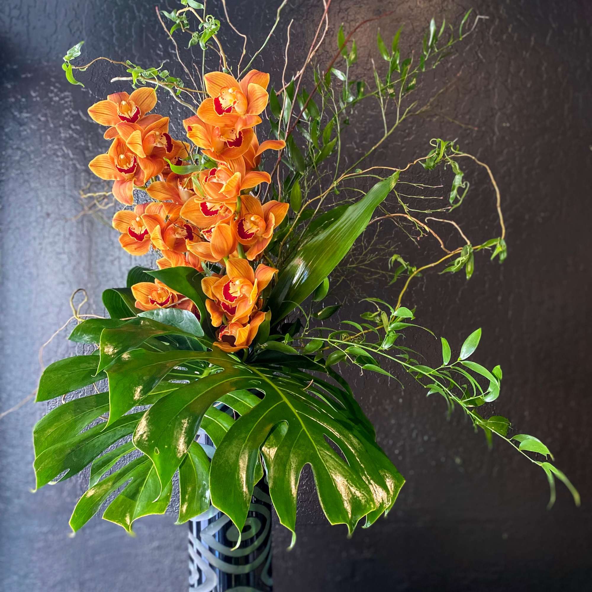 Tall arrangement of orange orchids with large monstera leaves in a patterned black cylinder vase