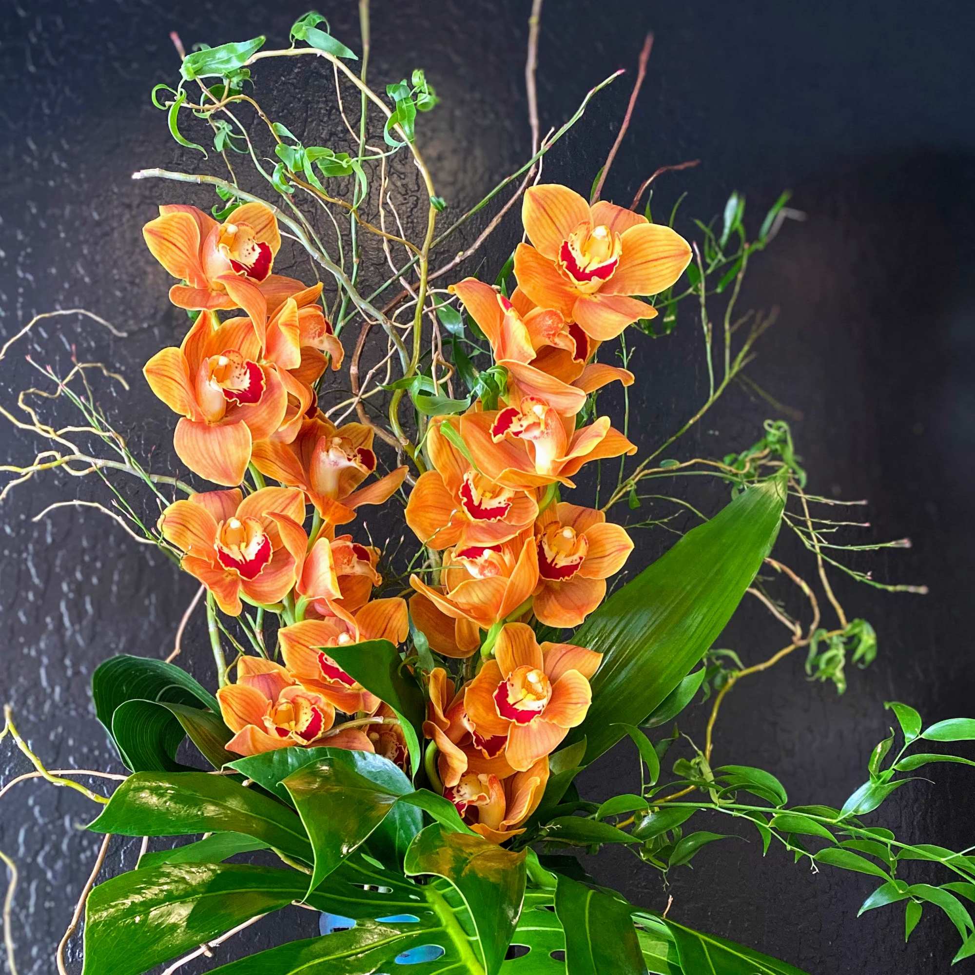 Tall arrangement of orange cymbidium orchids with curly branches and large tropical leaves