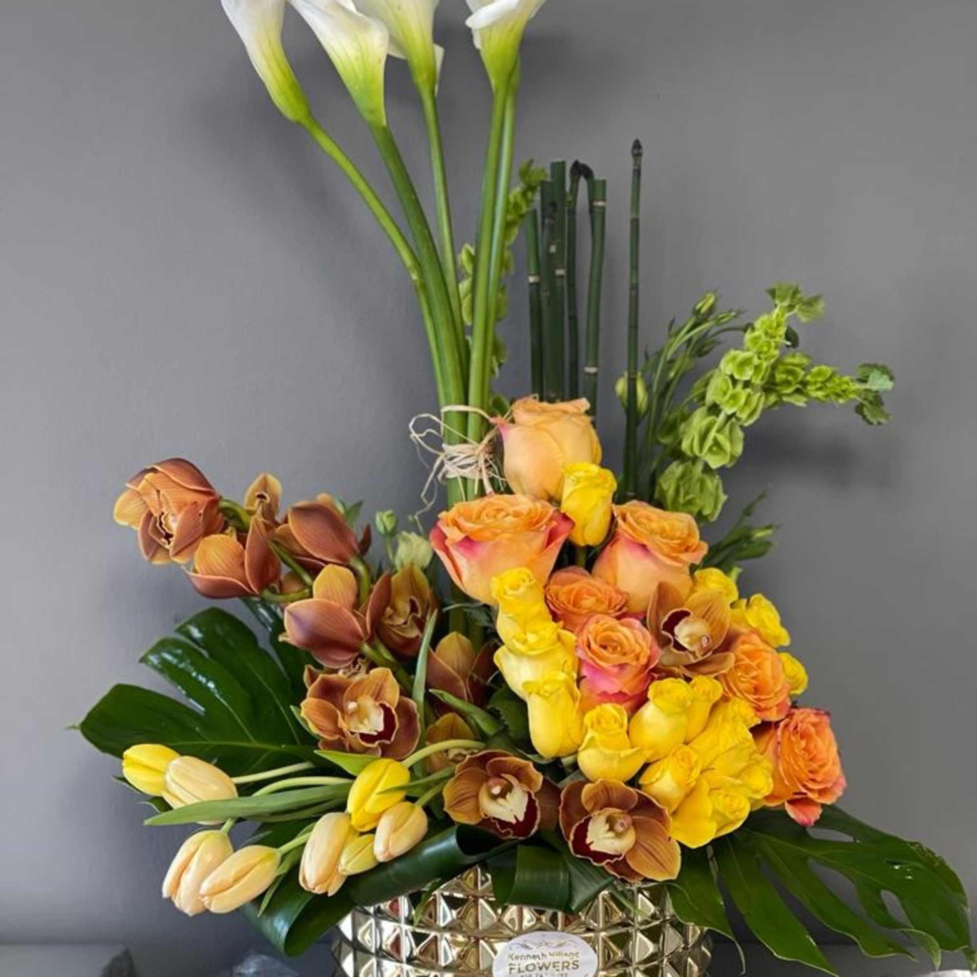 Tall floral arrangement with white calla lilies, roses, and orchids in a gold vase