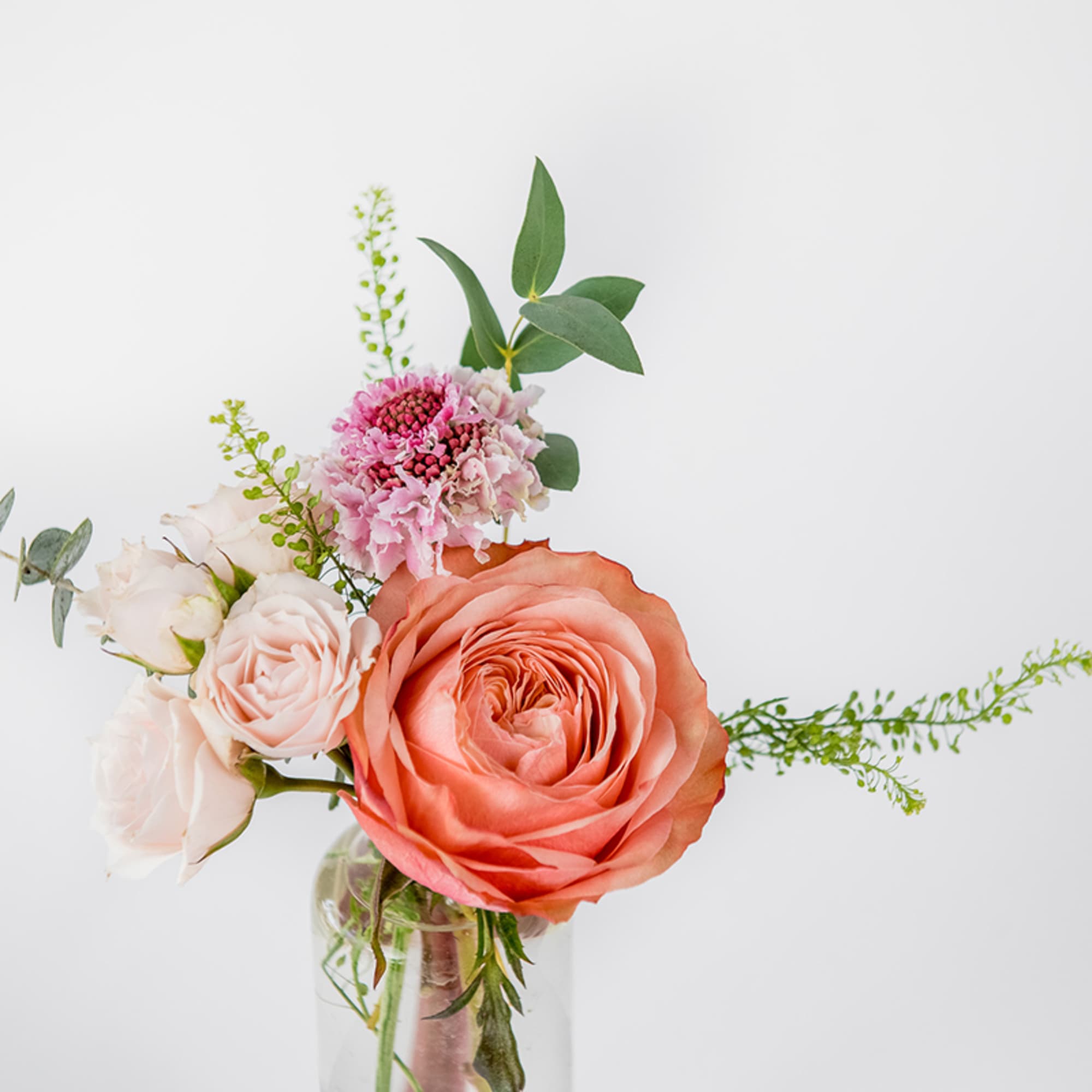 Small clear vase with a peach rose, blush roses, and a pink flower with airy greenery.