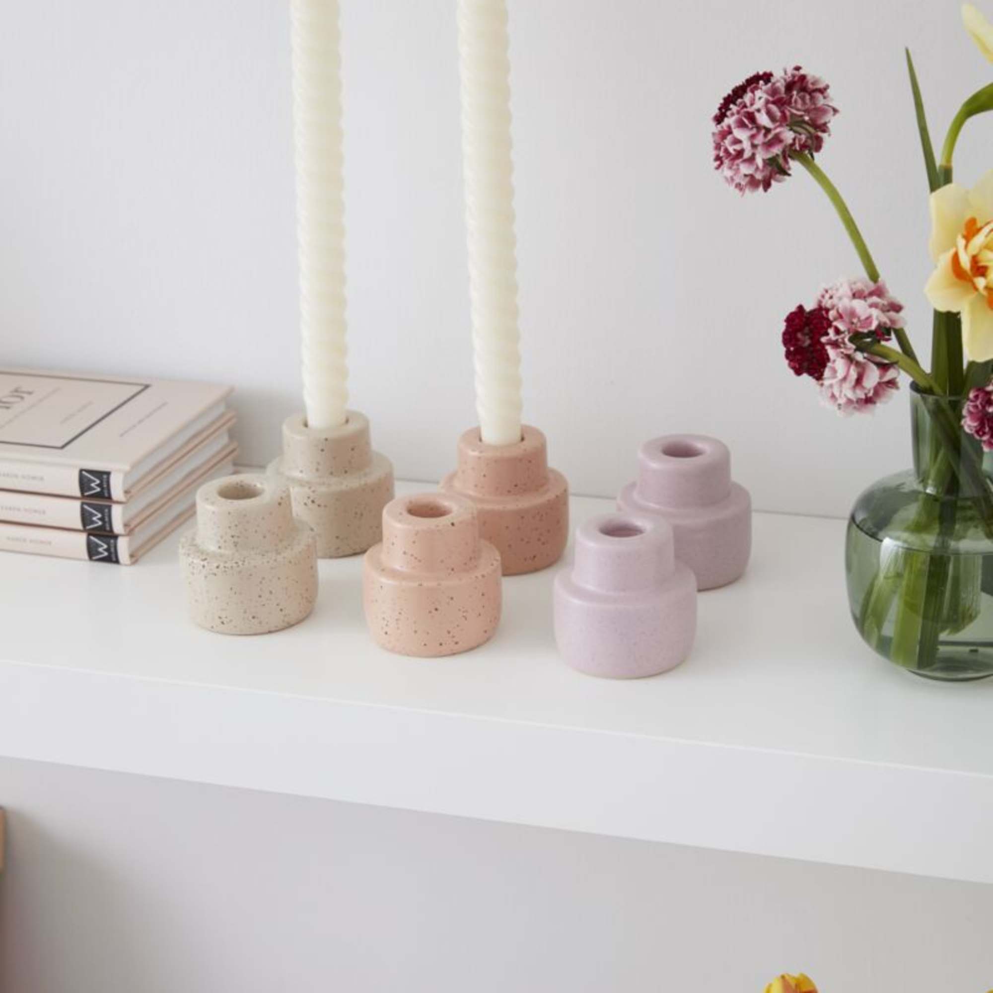 Pastel ceramic candle holders with white tapers on a shelf beside a small vase of mixed spring flowers