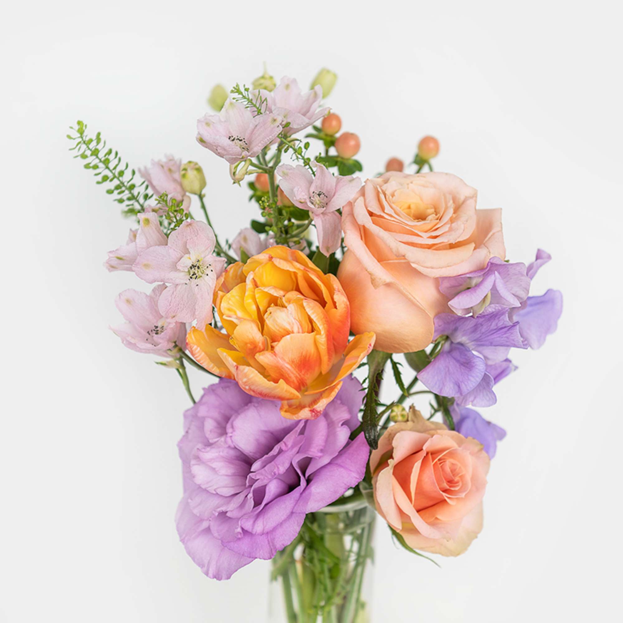 Small mixed bouquet of peach roses, yellow ranunculus, and purple flowers in a clear glass vase