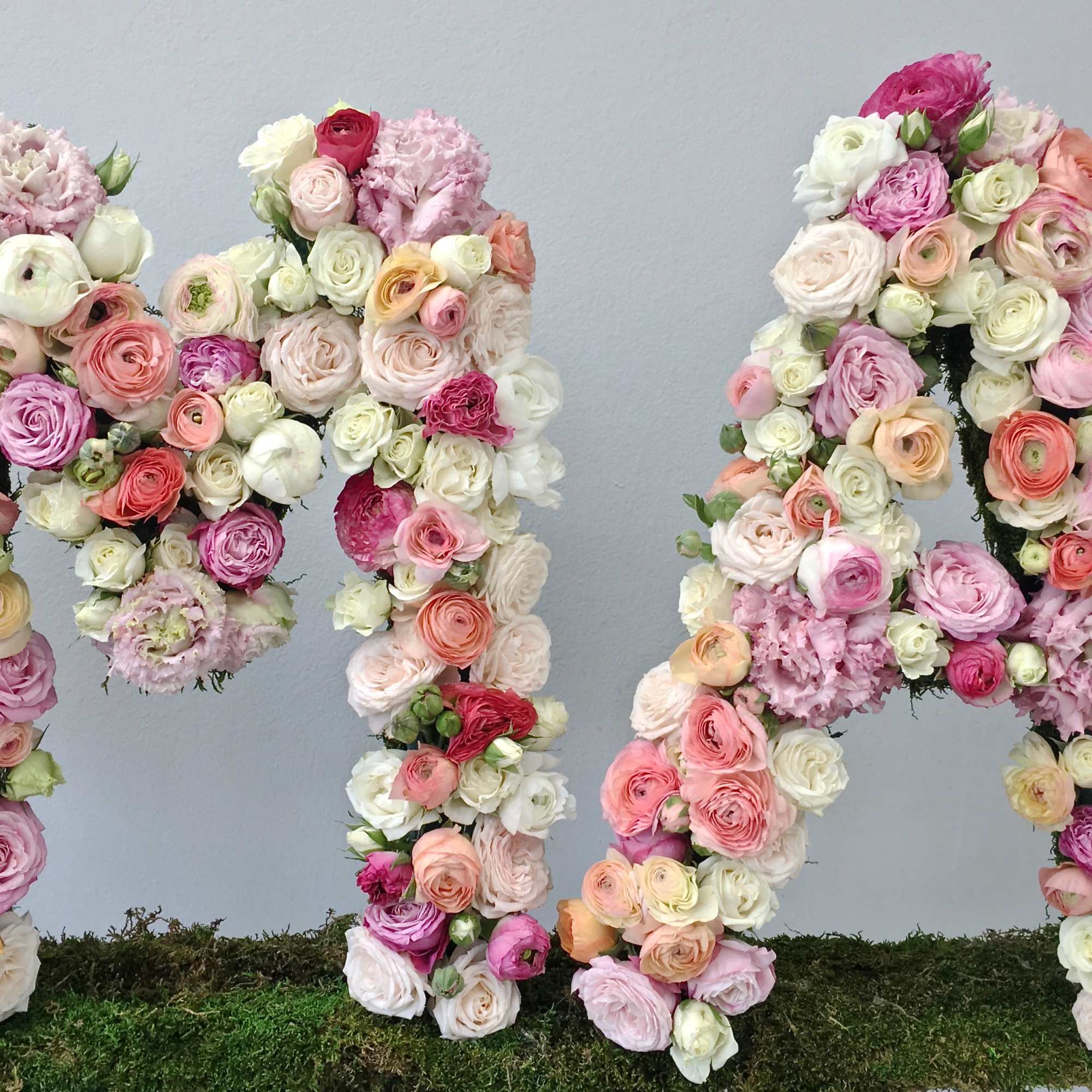 Large standing letters M and A made from pink, peach, and white roses and ranunculus on a moss base