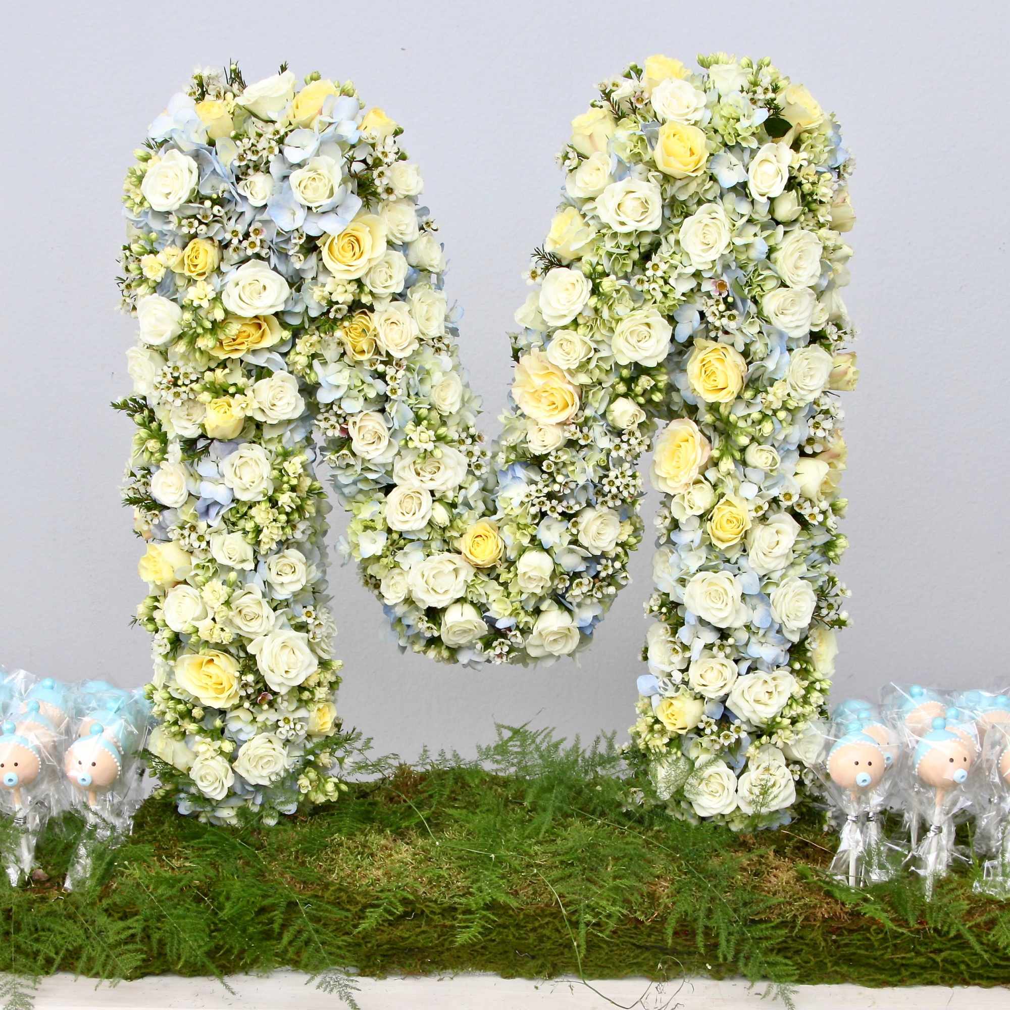 Large floral letter M in white, yellow, and pale blue flowers on a moss base with baby-themed cake pops.
