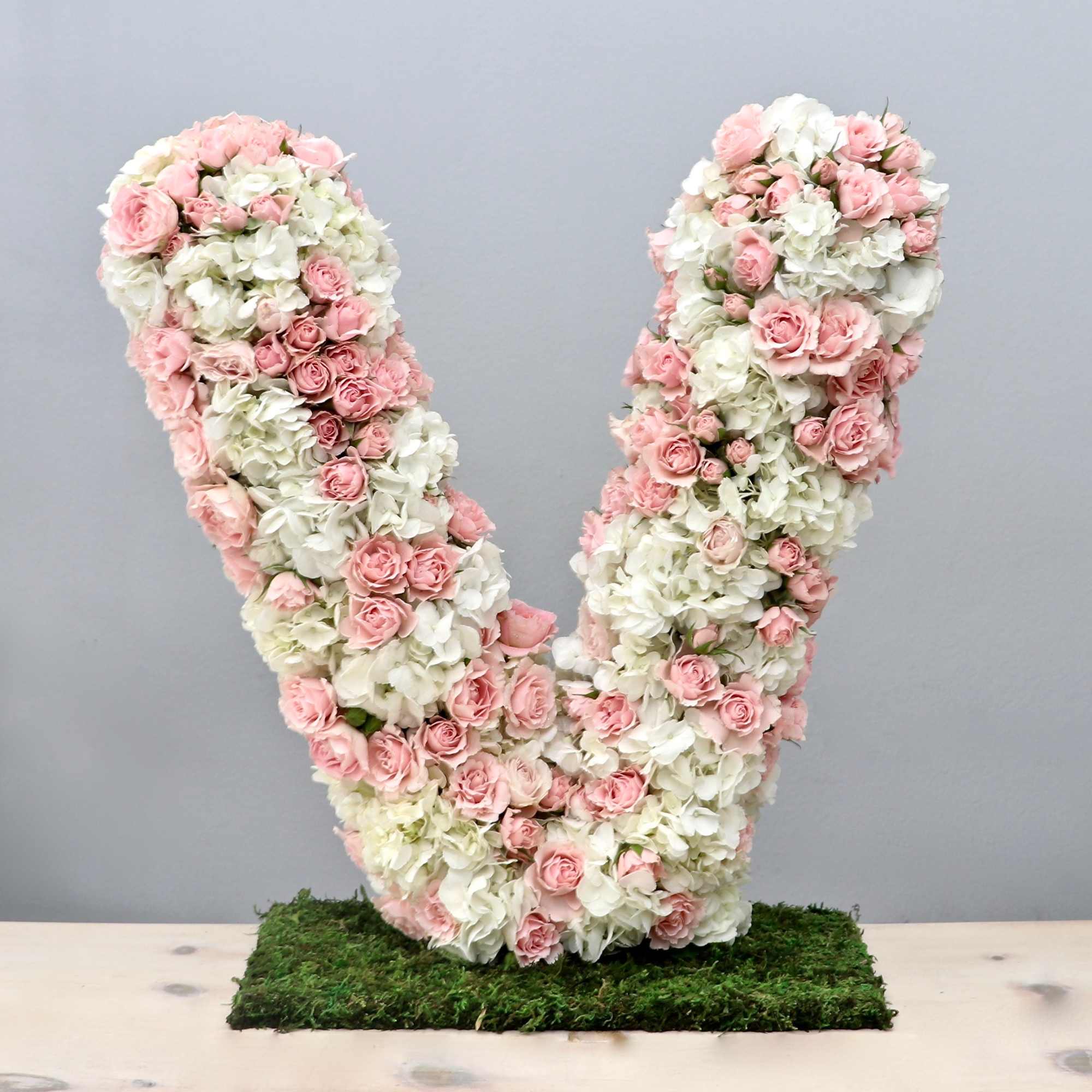 Large floral letter V made of pink roses and white hydrangeas on a moss-covered rectangular base