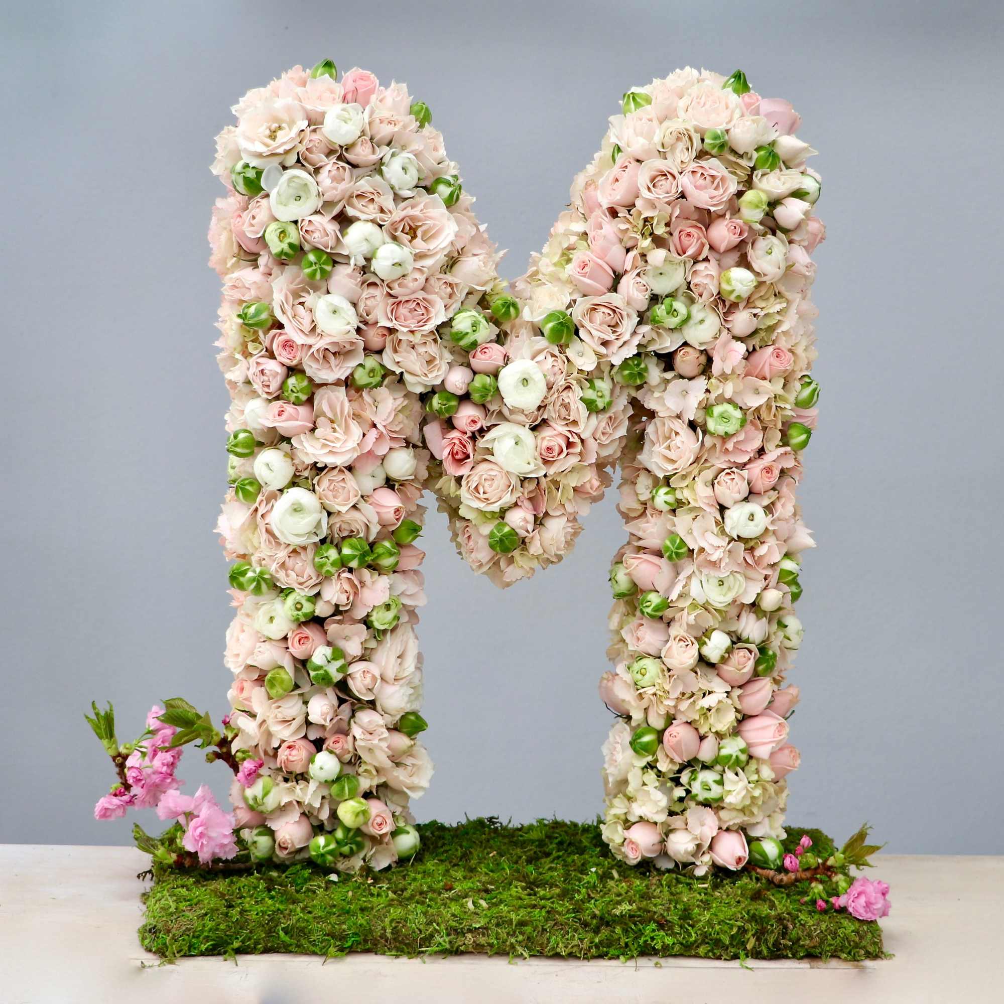 Large floral letter M made of blush pink and white blooms on a moss-covered base