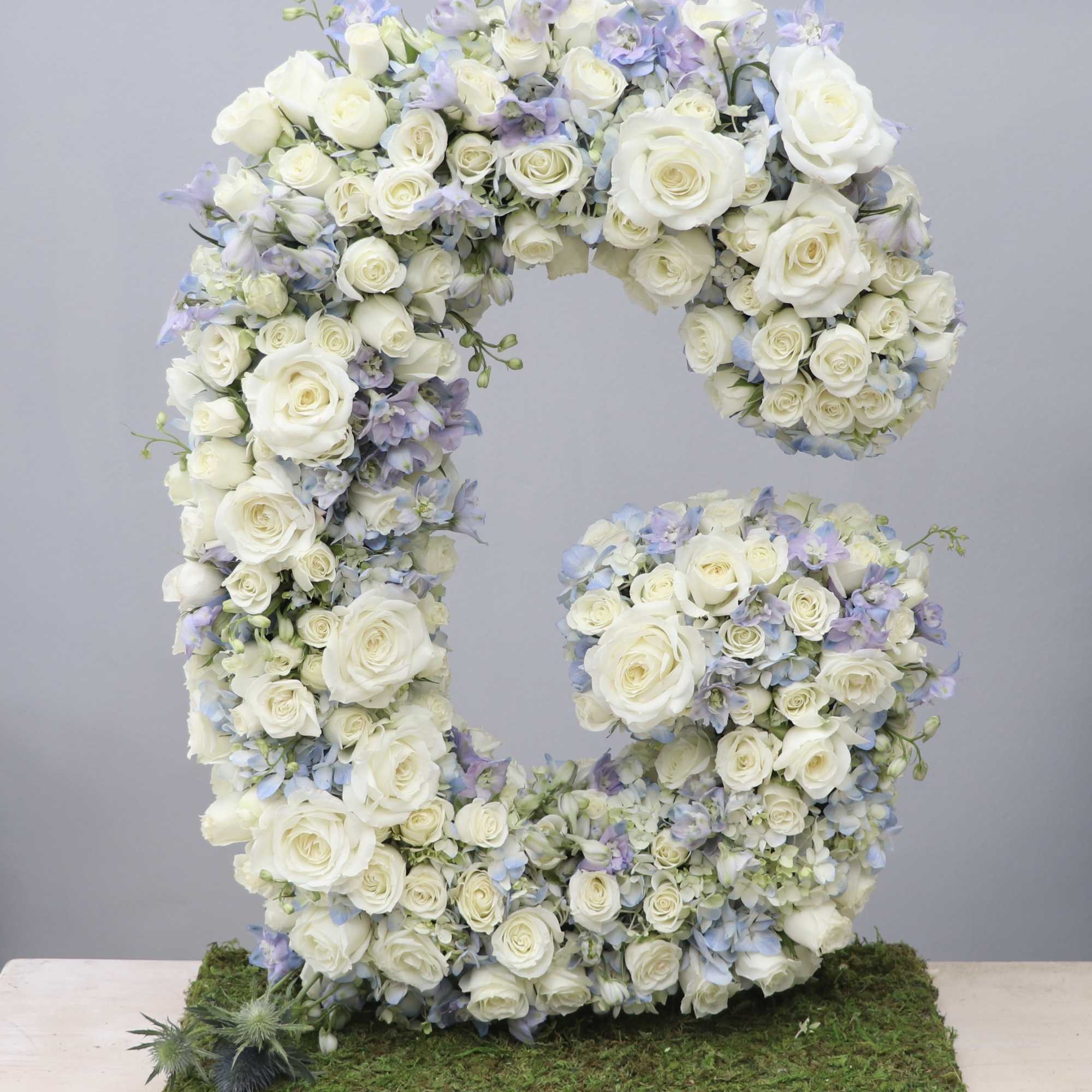 Tall floral letter G made of white roses and pale blue flowers on a moss-covered base