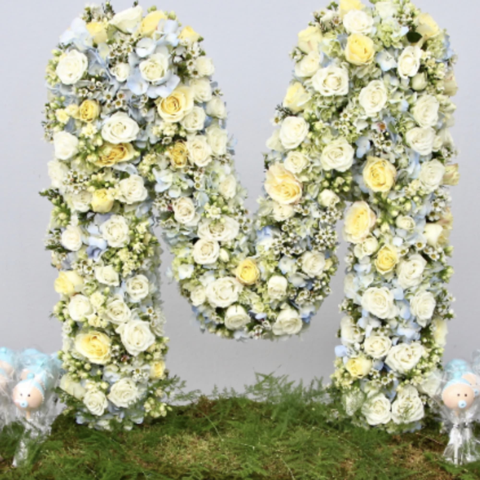 Large letter M tribute made of white, yellow, and blue flowers on a mossy base with small blue teddy bears.