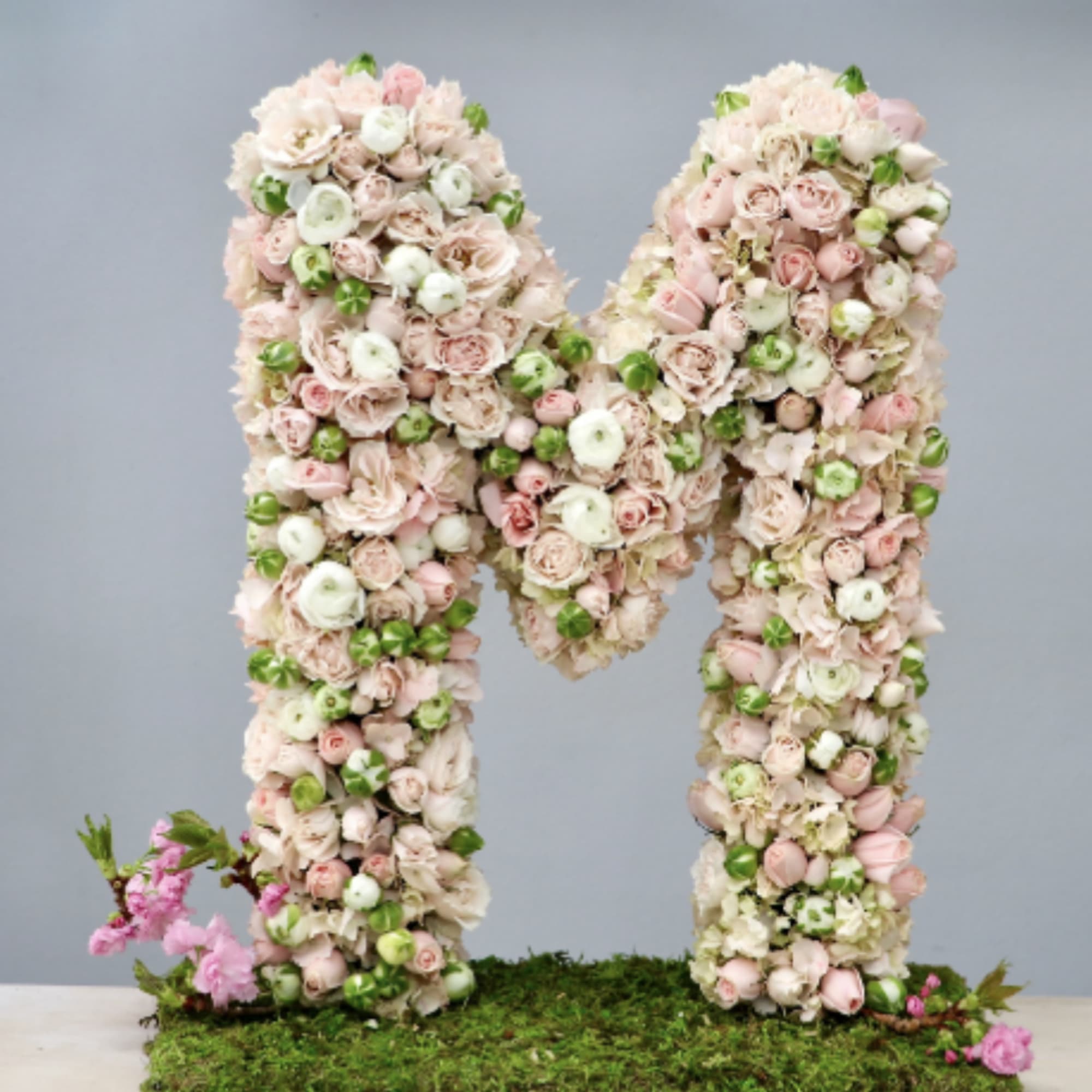 Tall floral letter M made of pastel pink and white blooms on a moss base
