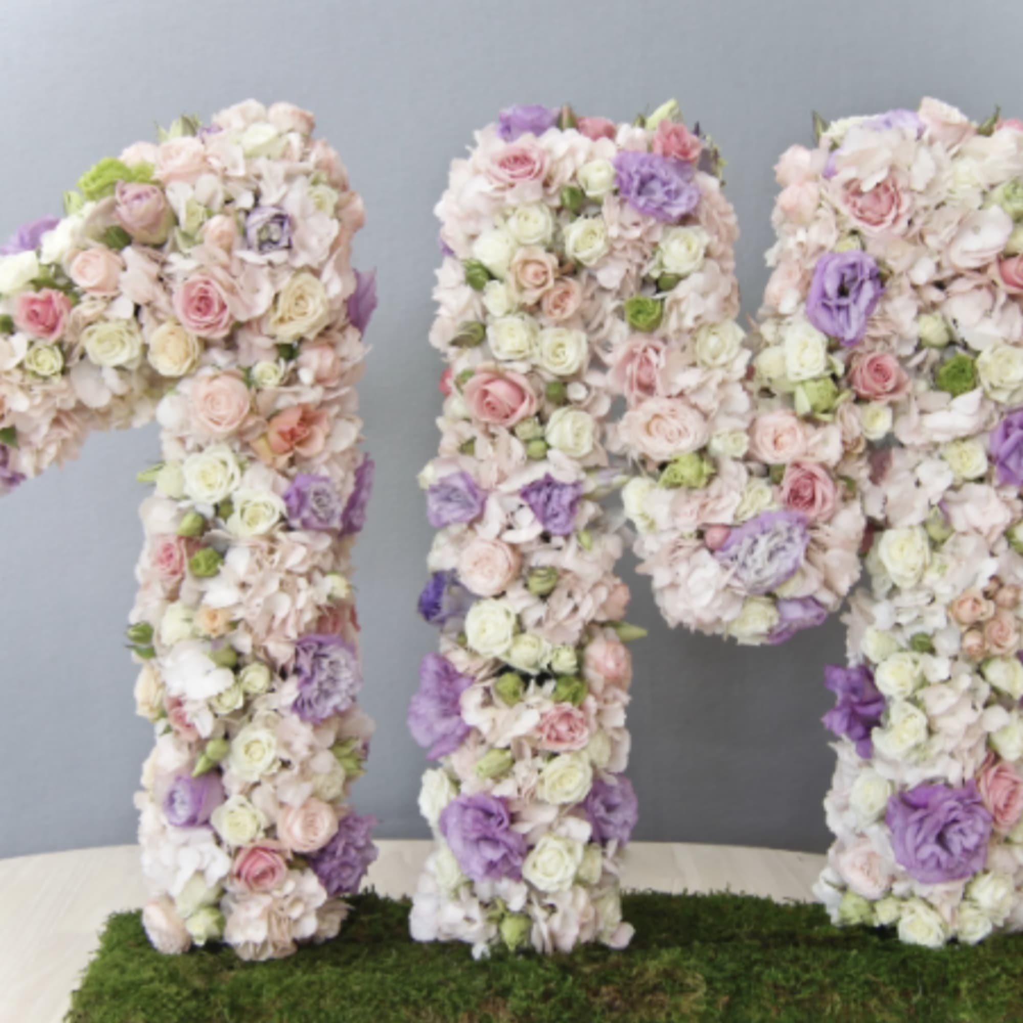 Large floral sculpture of the characters 1M in pastel pink, white, and lavender flowers on a moss base