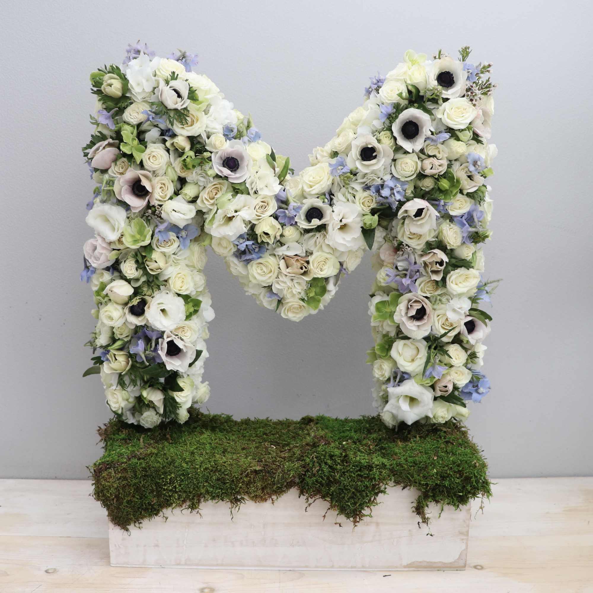 Floral letter M made of white and pastel flowers on a moss-covered rectangular base