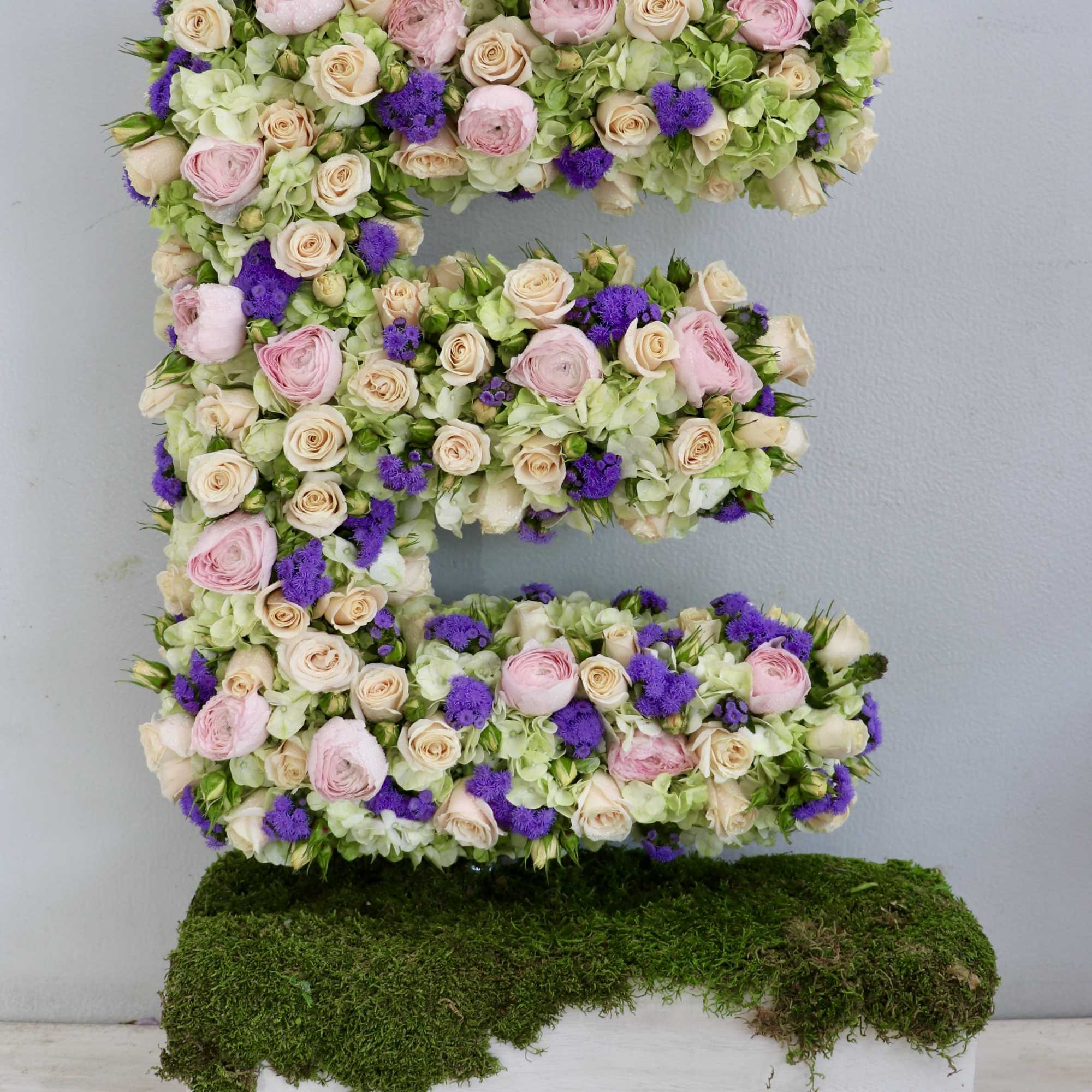Floral letter E made of pastel roses, hydrangeas, and purple blooms on a moss-topped white box base