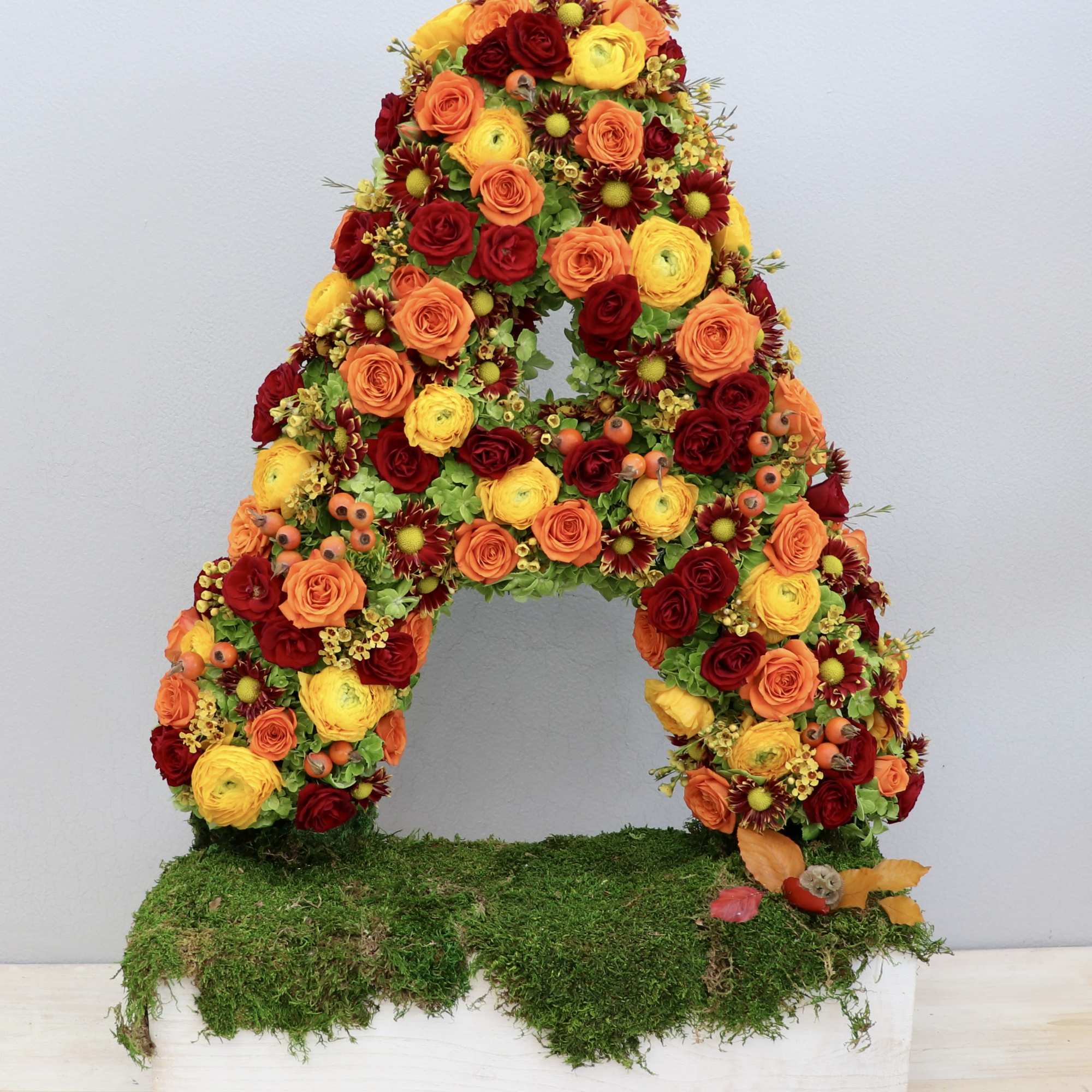 Tall letter A floral arrangement in warm red, orange, and yellow flowers on a moss-covered white base