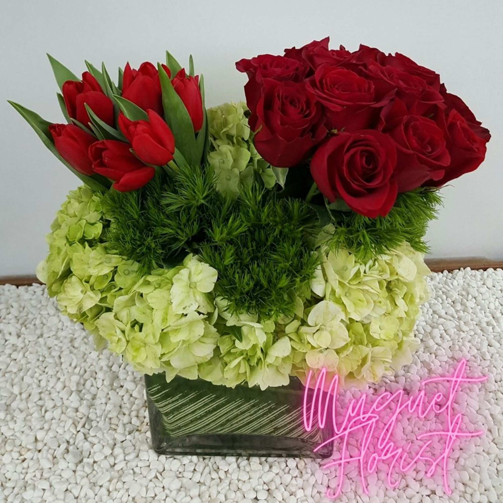 Red roses and tulips arranged with pale green hydrangeas in a square vase.