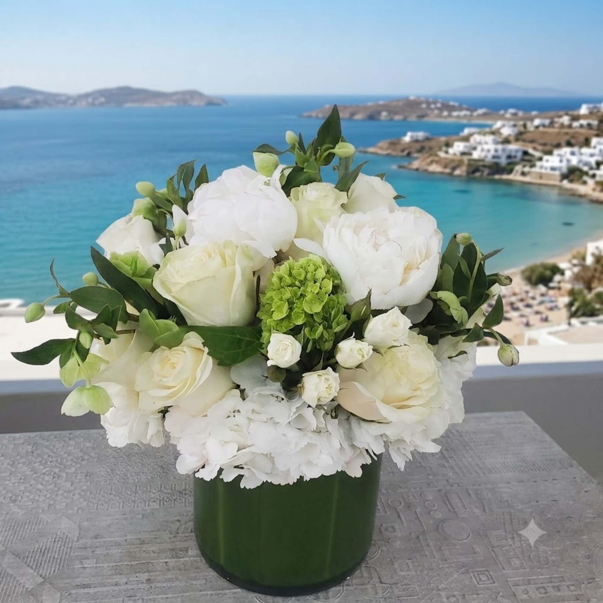 White floral arrangement in a dark green vase with a seaside view behind it