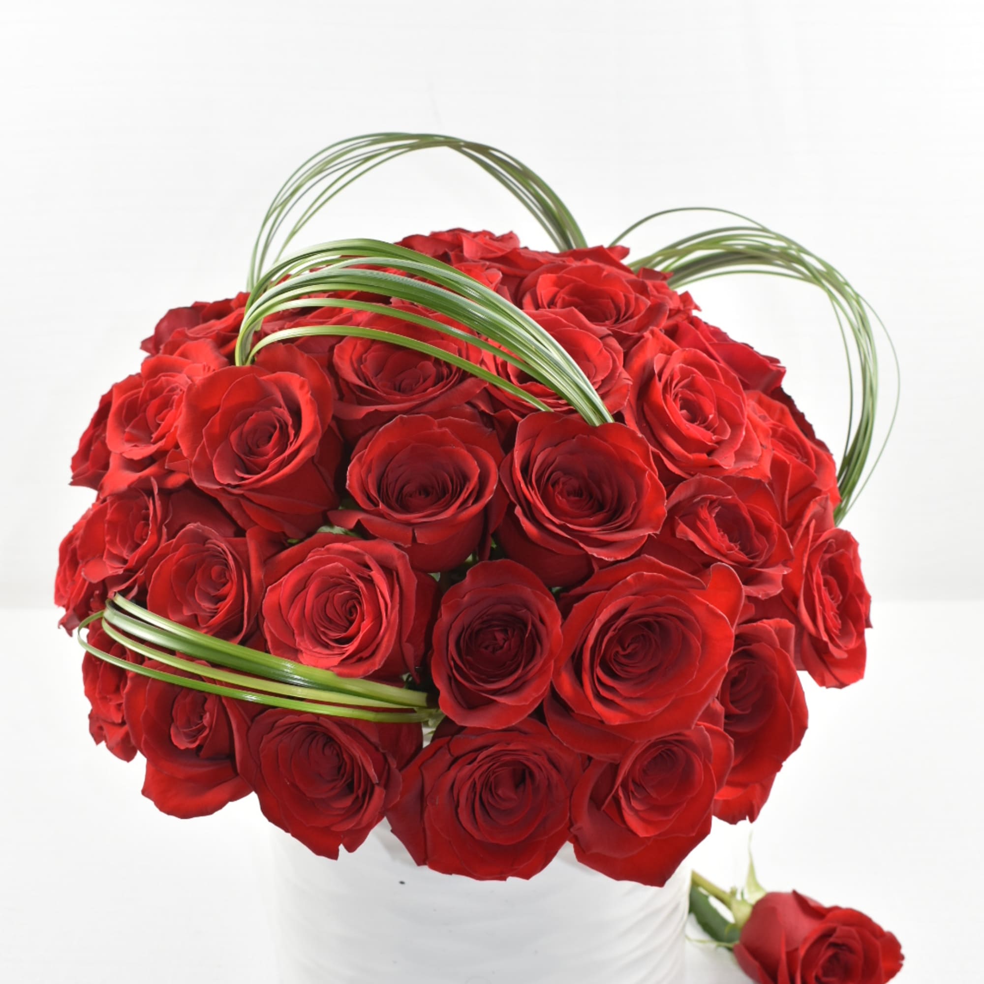 Compact arrangement of red roses with looped green blades in a white ceramic vase.