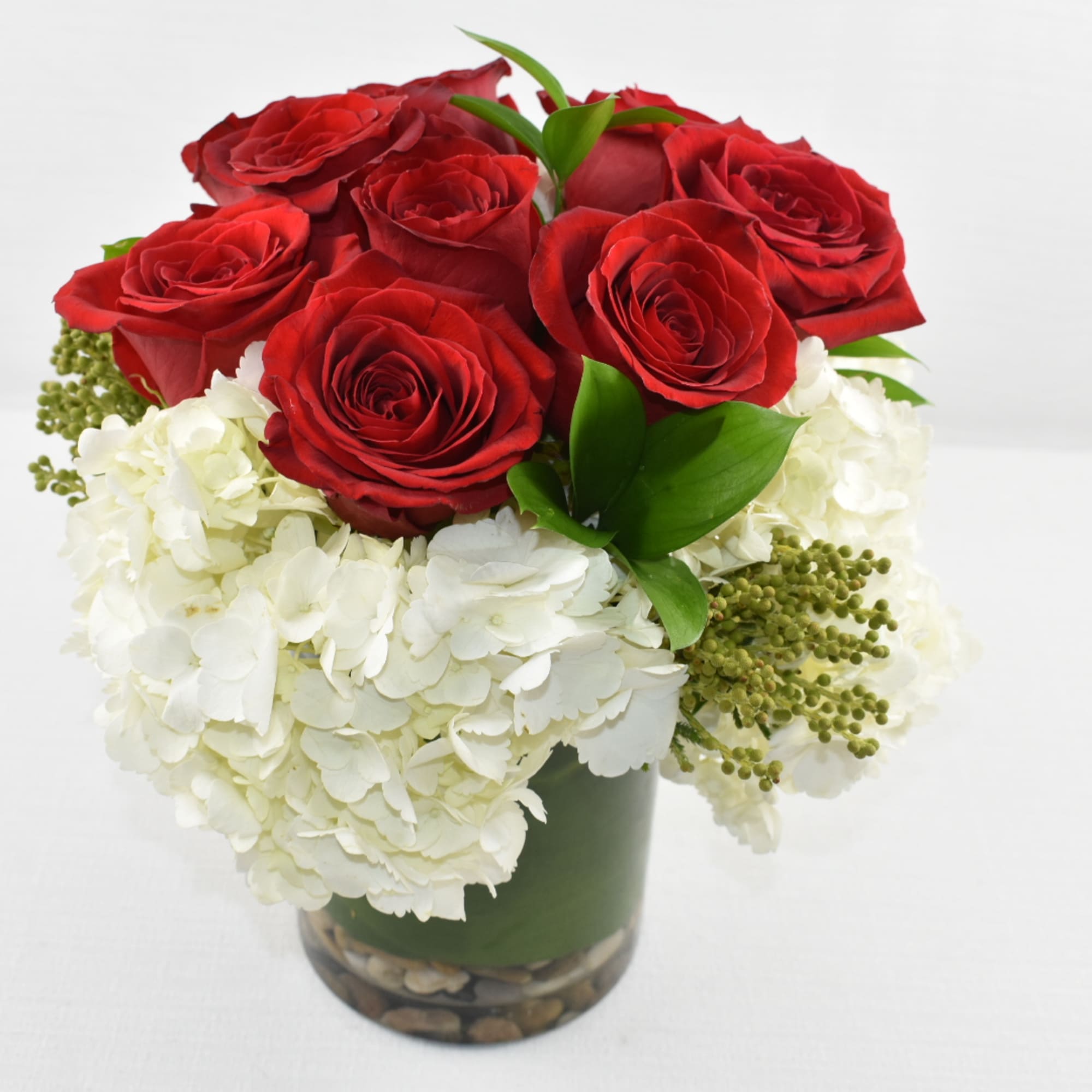 Compact arrangement of red roses and white hydrangeas in a glass cylinder vase lined with green and stones