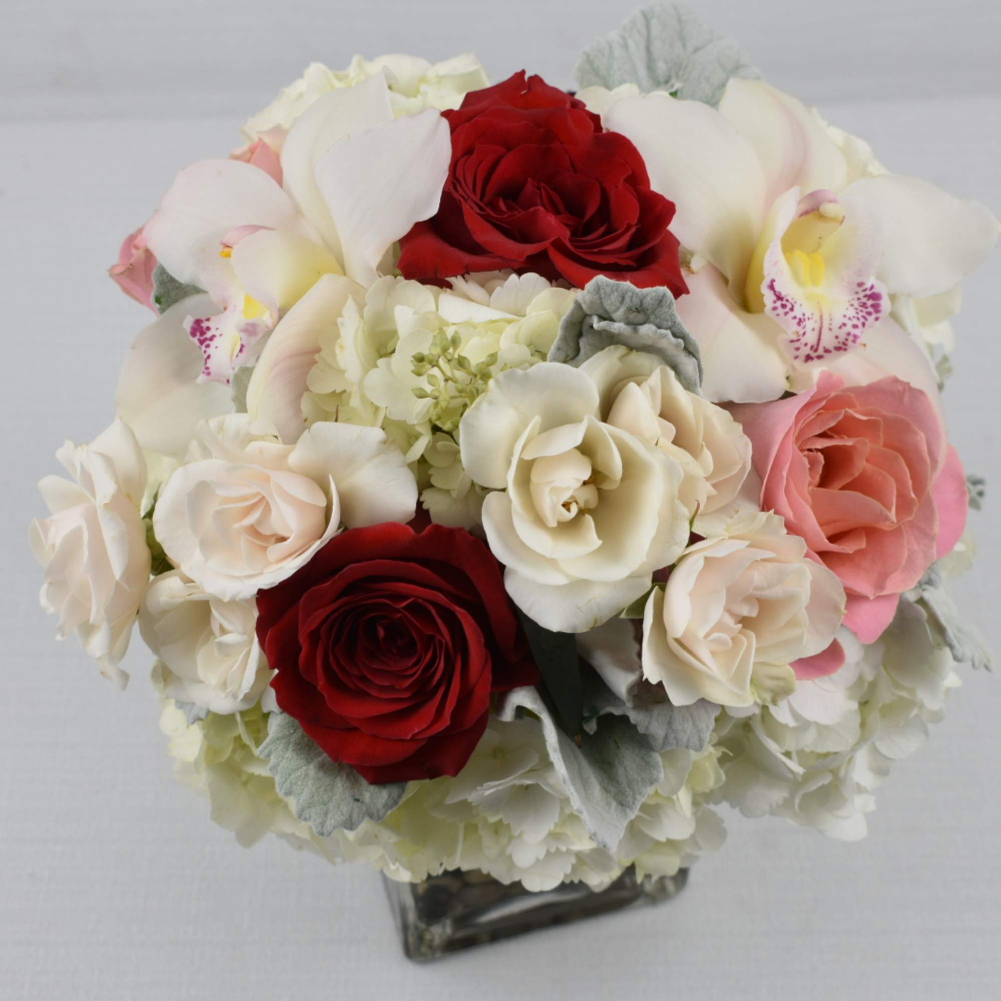 Compact arrangement of red, white, and pink roses with orchids in a clear glass cube vase