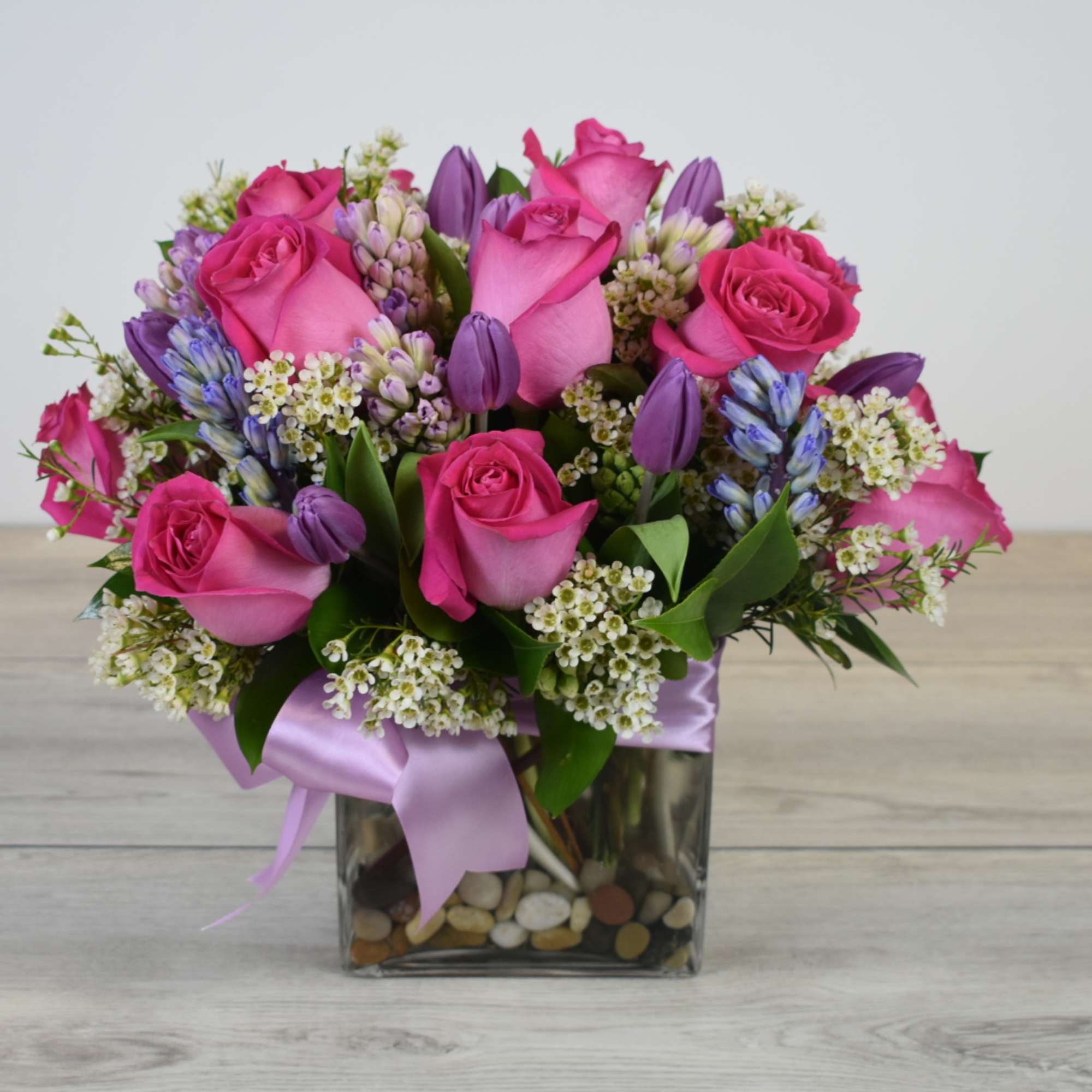 Compact arrangement of pink roses with purple tulips and hyacinths in a glass cube vase with ribbon.