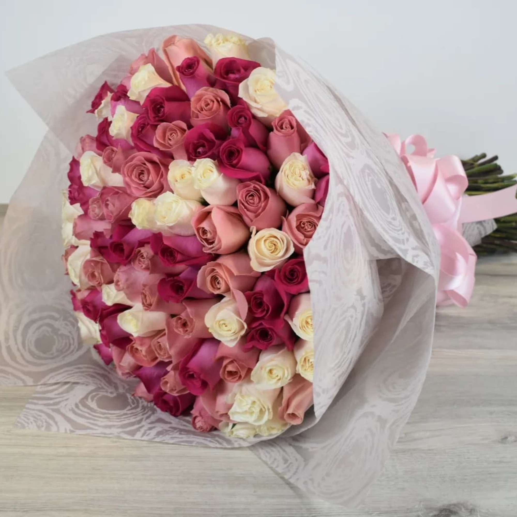 Large hand-tied bouquet of pink and cream roses wrapped in patterned white paper with a pink ribbon.