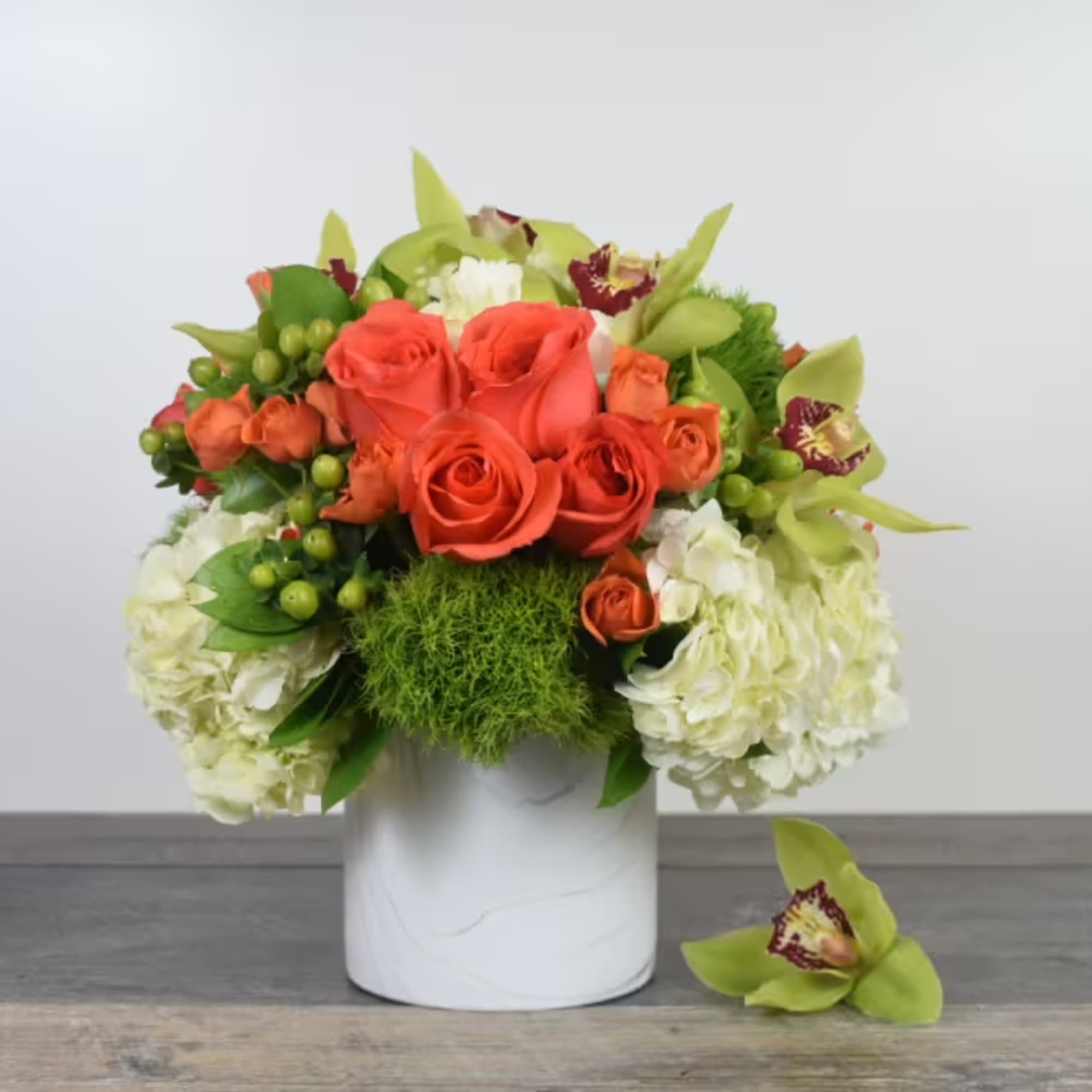 Compact arrangement of orange roses, green orchids, and white hydrangeas in a white cylindrical vase