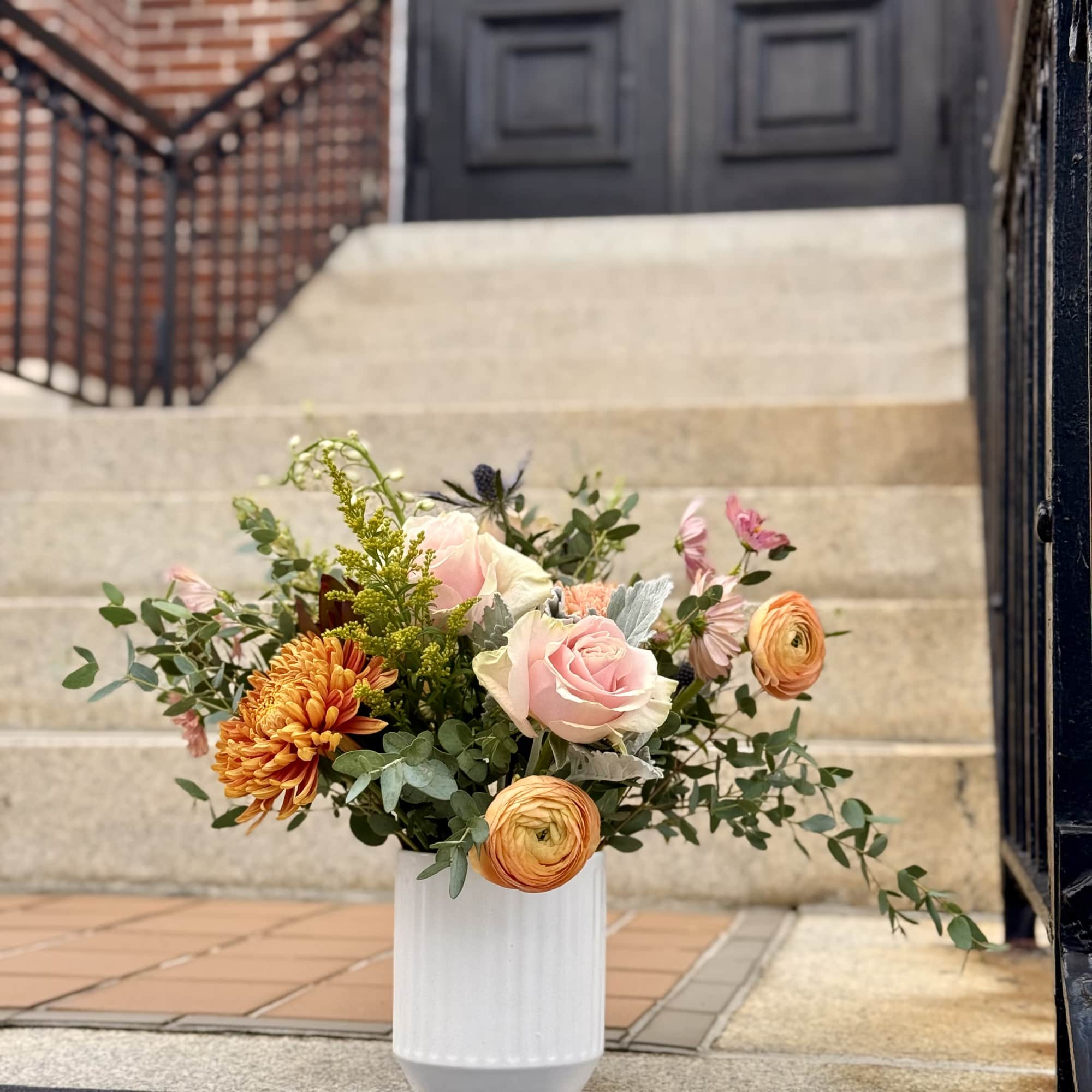 Arrangement of blush roses, orange chrysanthemums, and ranunculus in a white vase on outdoor steps