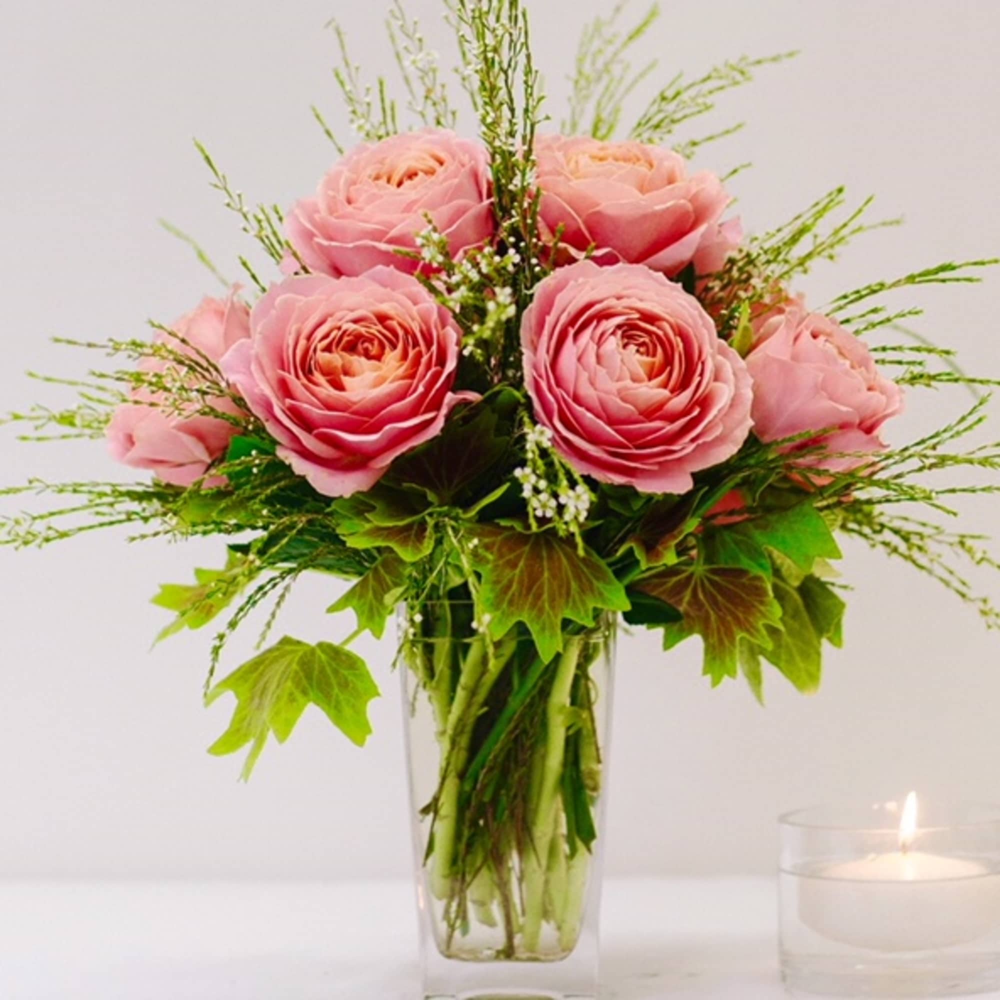 Pink roses arranged in a clear glass vase with greenery and a lit candle nearby