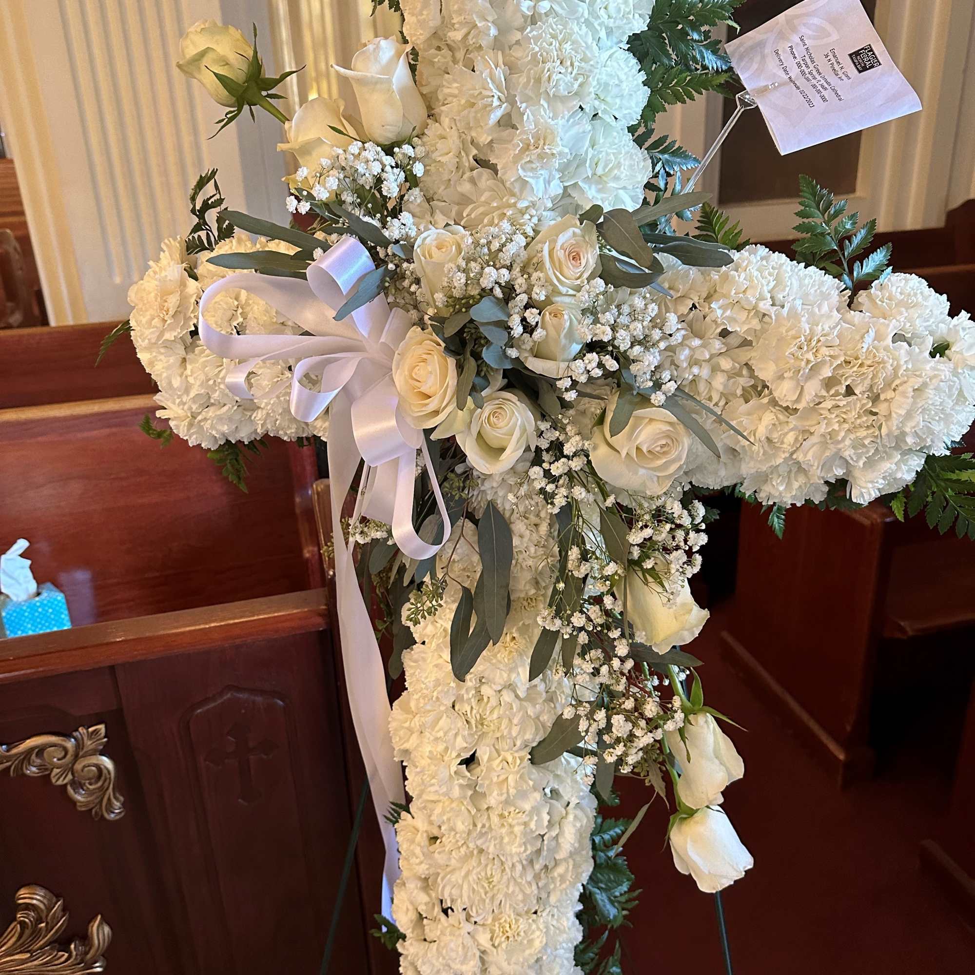 Standing white floral cross with roses, carnations, and ribbon displayed on an easel.