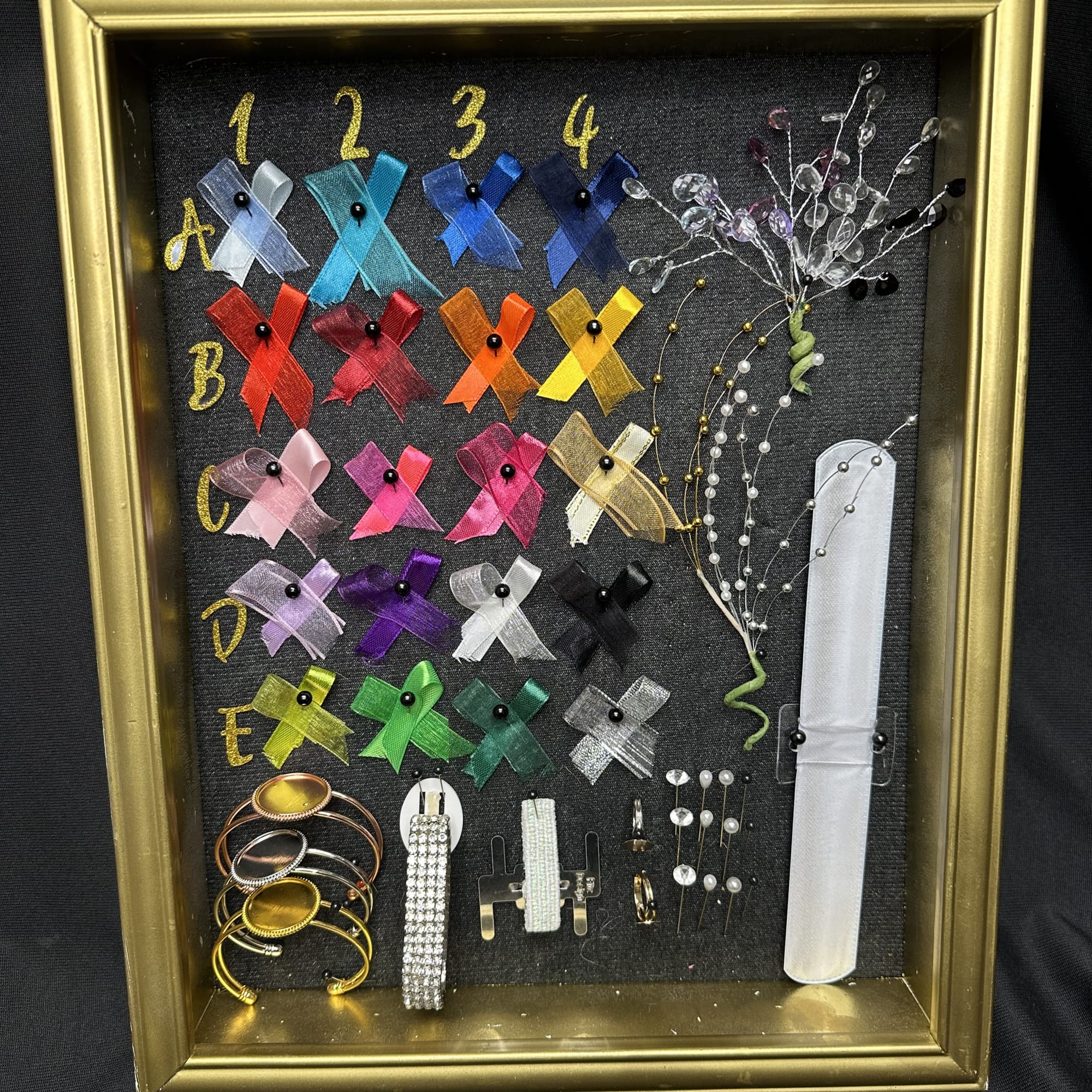 Framed display of colorful ribbon pins and jewelry pieces on black fabric