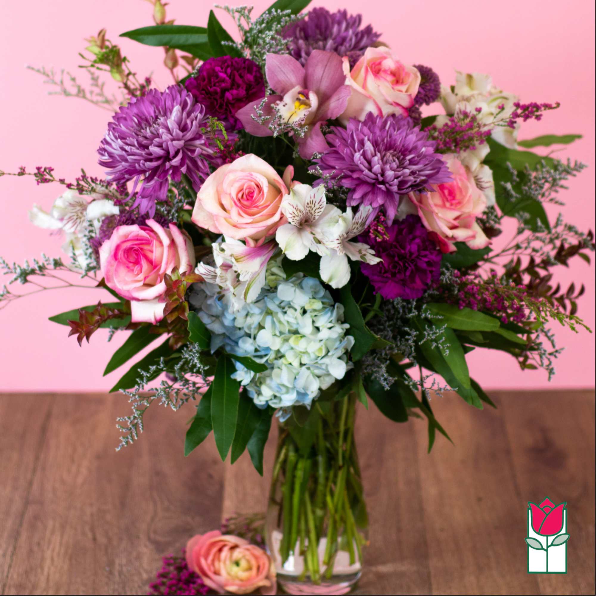 Mixed bouquet of pink roses, purple mums, orchids, and hydrangea in a clear glass vase on a wooden table.