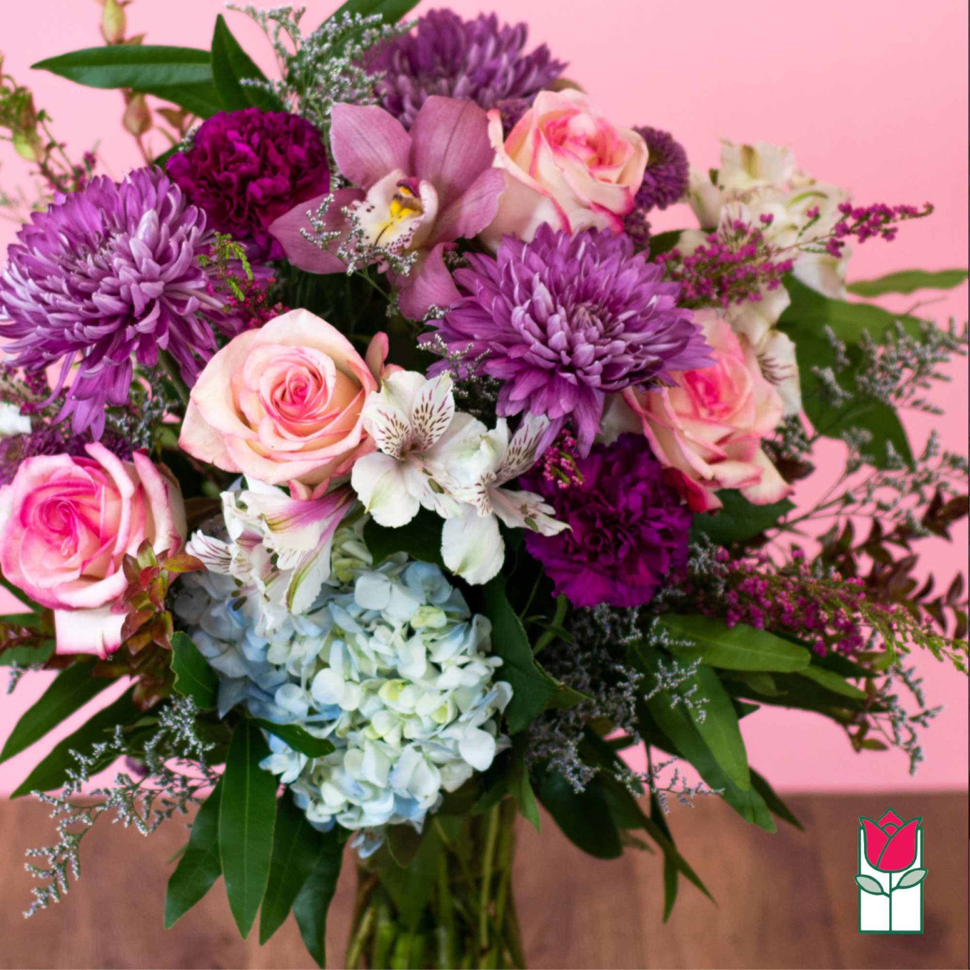Mixed bouquet of pink roses, purple mums, white alstroemeria, and blue hydrangea in a clear glass vase