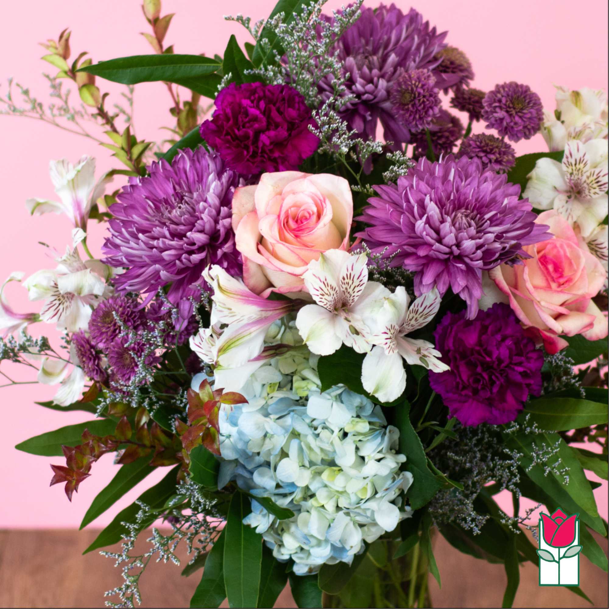 Mixed bouquet of purple mums, pink roses, white alstroemeria, and blue hydrangea in a glass vase