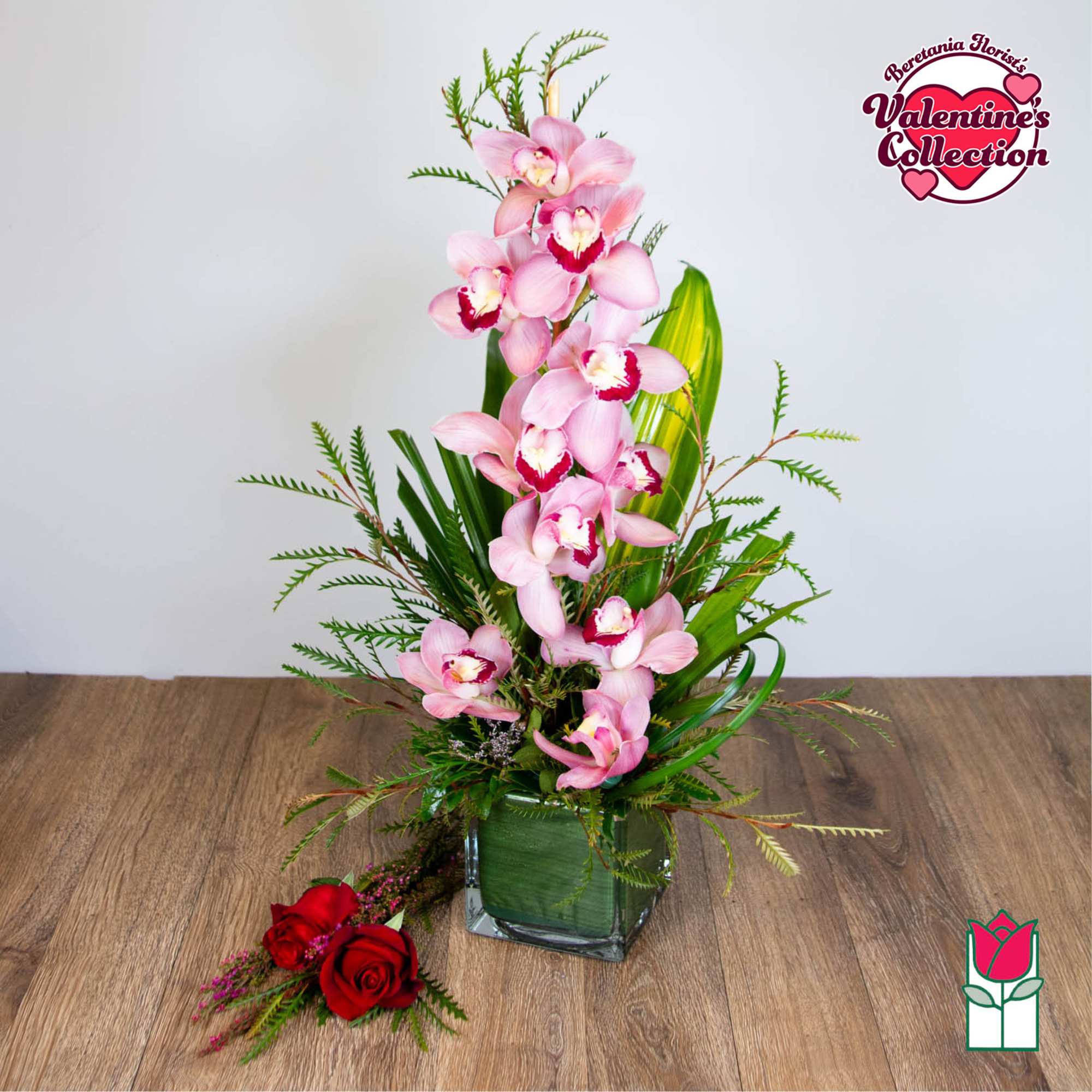 Tall pink cymbidium orchid arrangement in a glass cube vase with greenery and two red roses at the base.