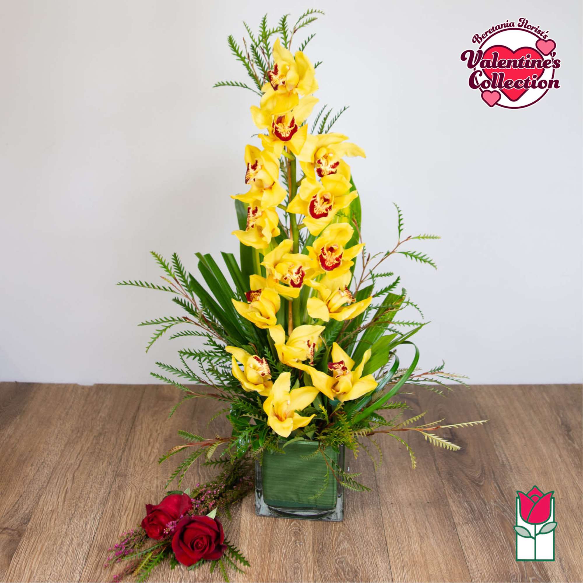 Tall yellow cymbidium orchid arrangement in a glass cube vase with loose red roses on the table
