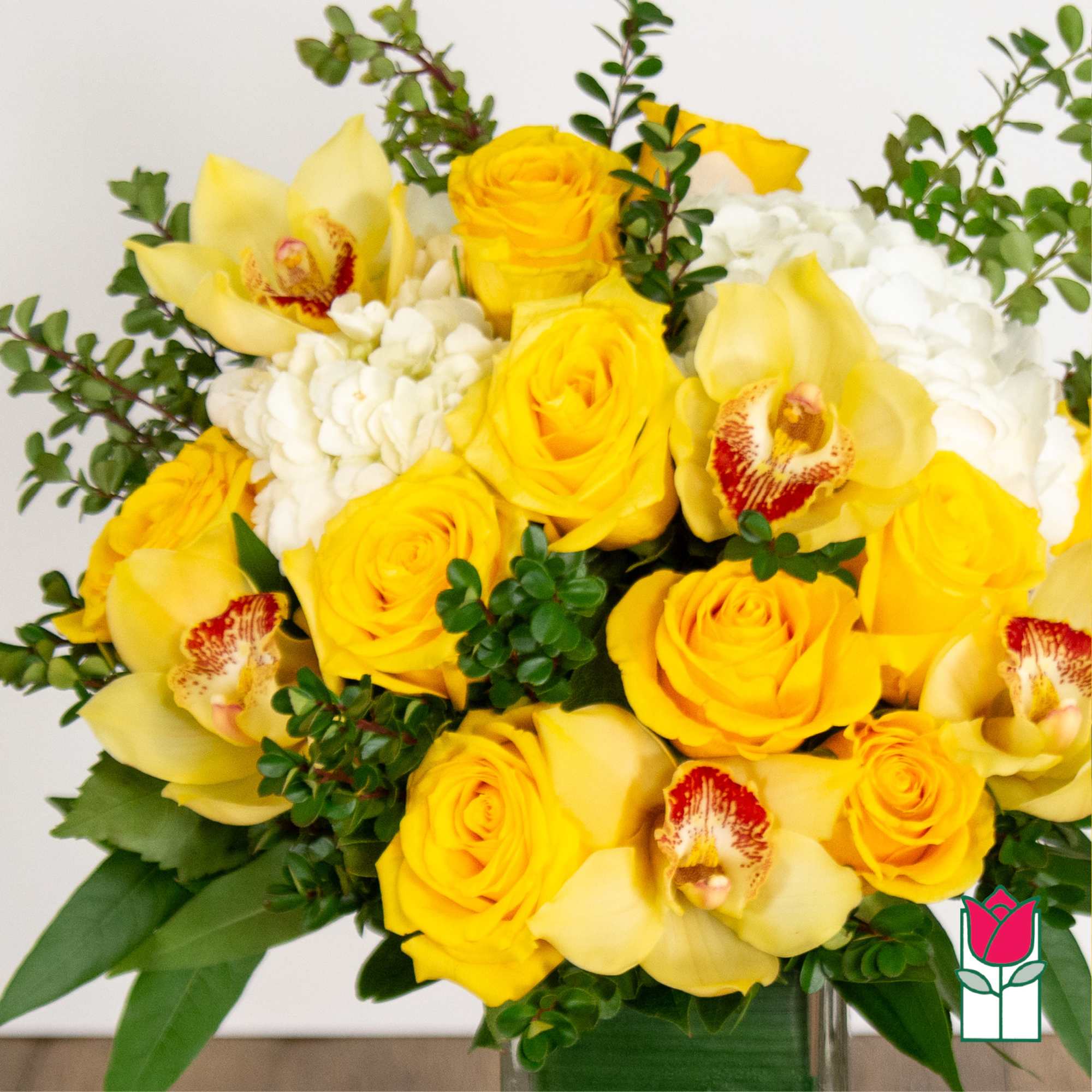 Yellow roses and orchids arranged in a vase with white blooms and greenery.