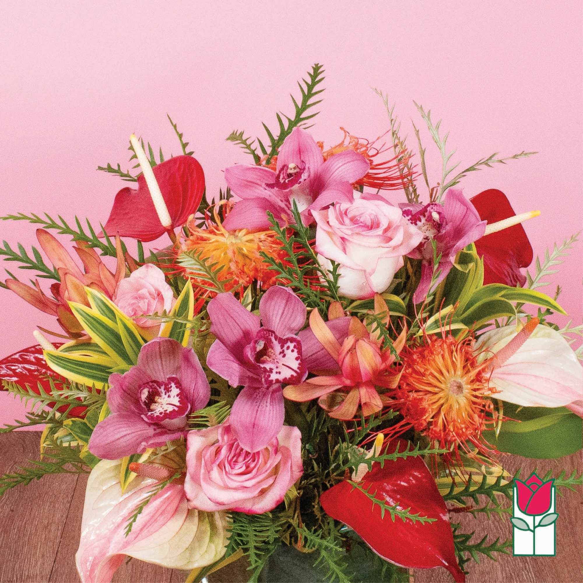 Vibrant arrangement of pink roses, orchids, and tropical blooms in a clear glass vase against a pink background.