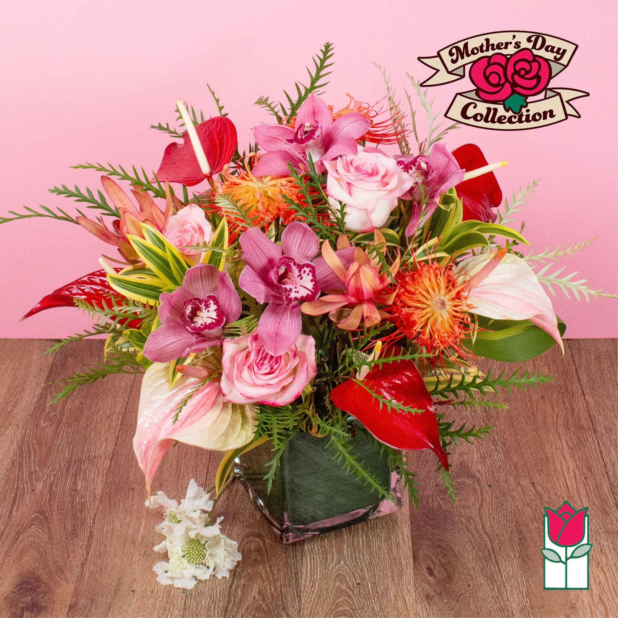 Vibrant pink orchids, roses, and tropical blooms in a clear glass cube vase on a wood surface.