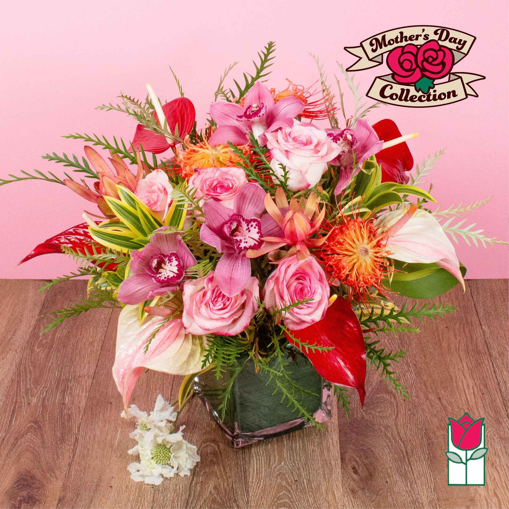 Mixed bouquet of pink roses, orchids, and tropical blooms in a square glass vase against a pink background.