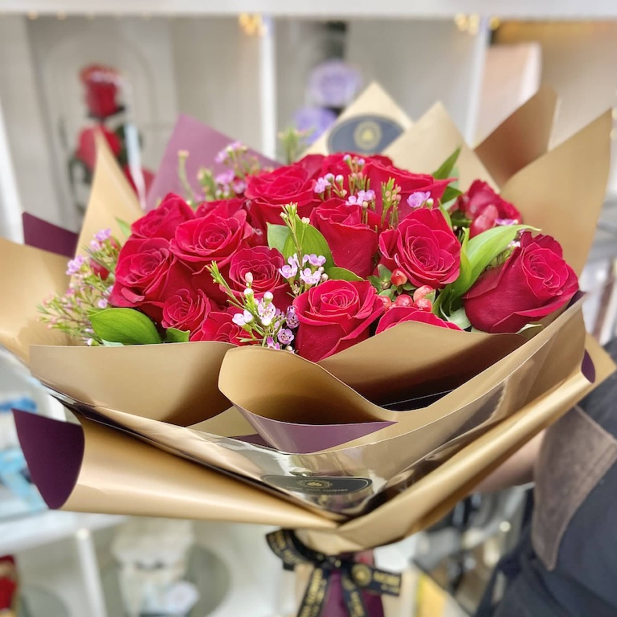 Handheld bouquet of red roses with small pink blooms wrapped in gold and plum paper.