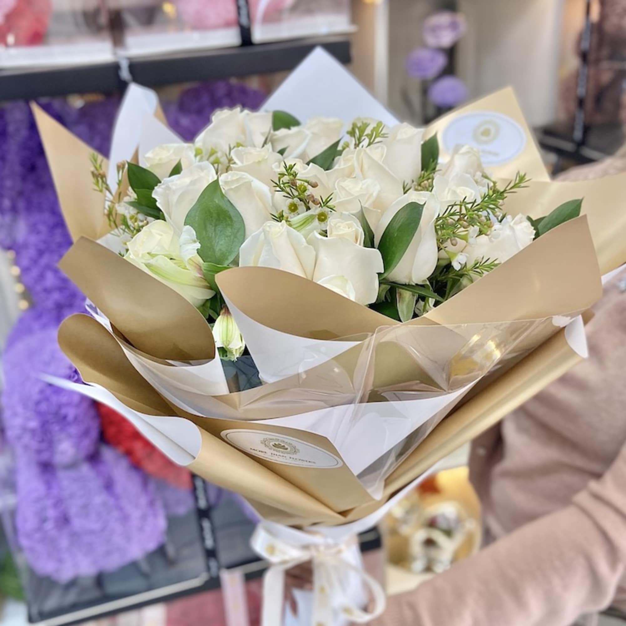 Hand-tied bouquet of white roses wrapped in gold and white paper with ribbon, held in a shop.