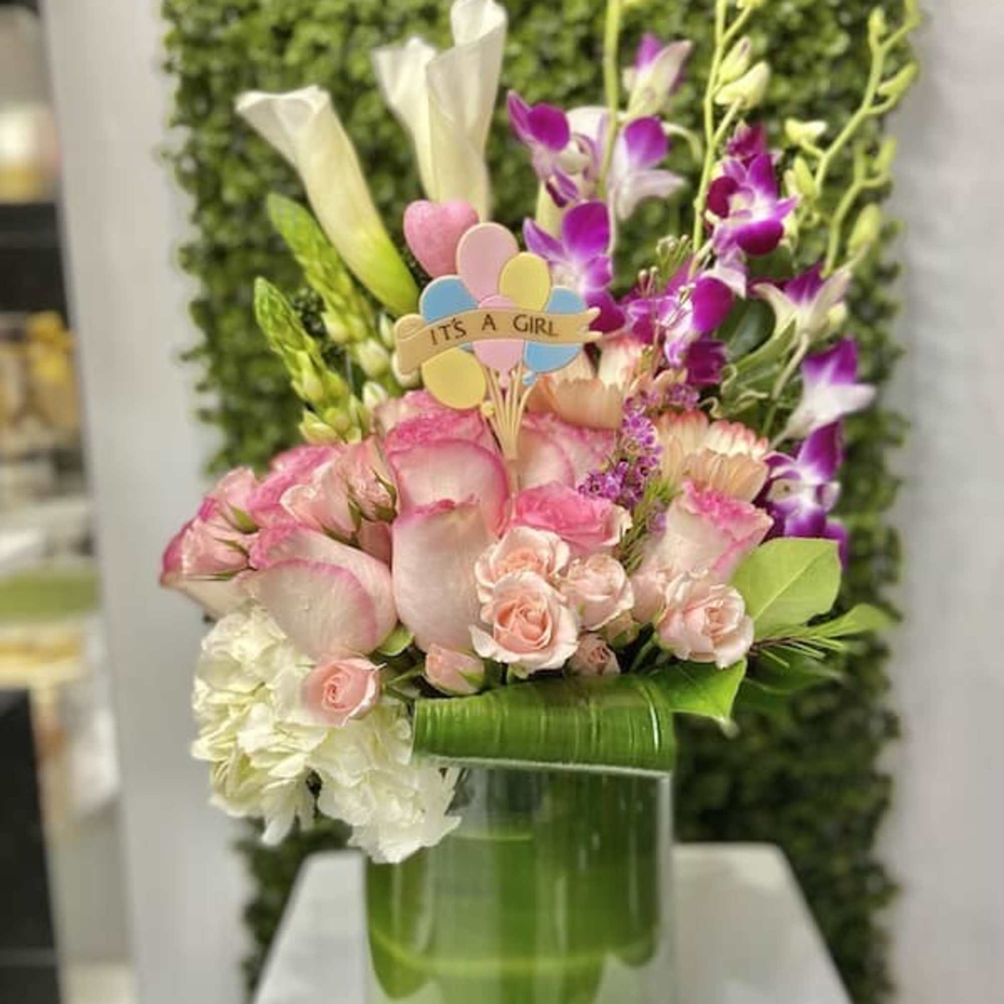 Pink and white flower arrangement with an It's a Girl pick in a glass cylinder vase lined with green leaves.
