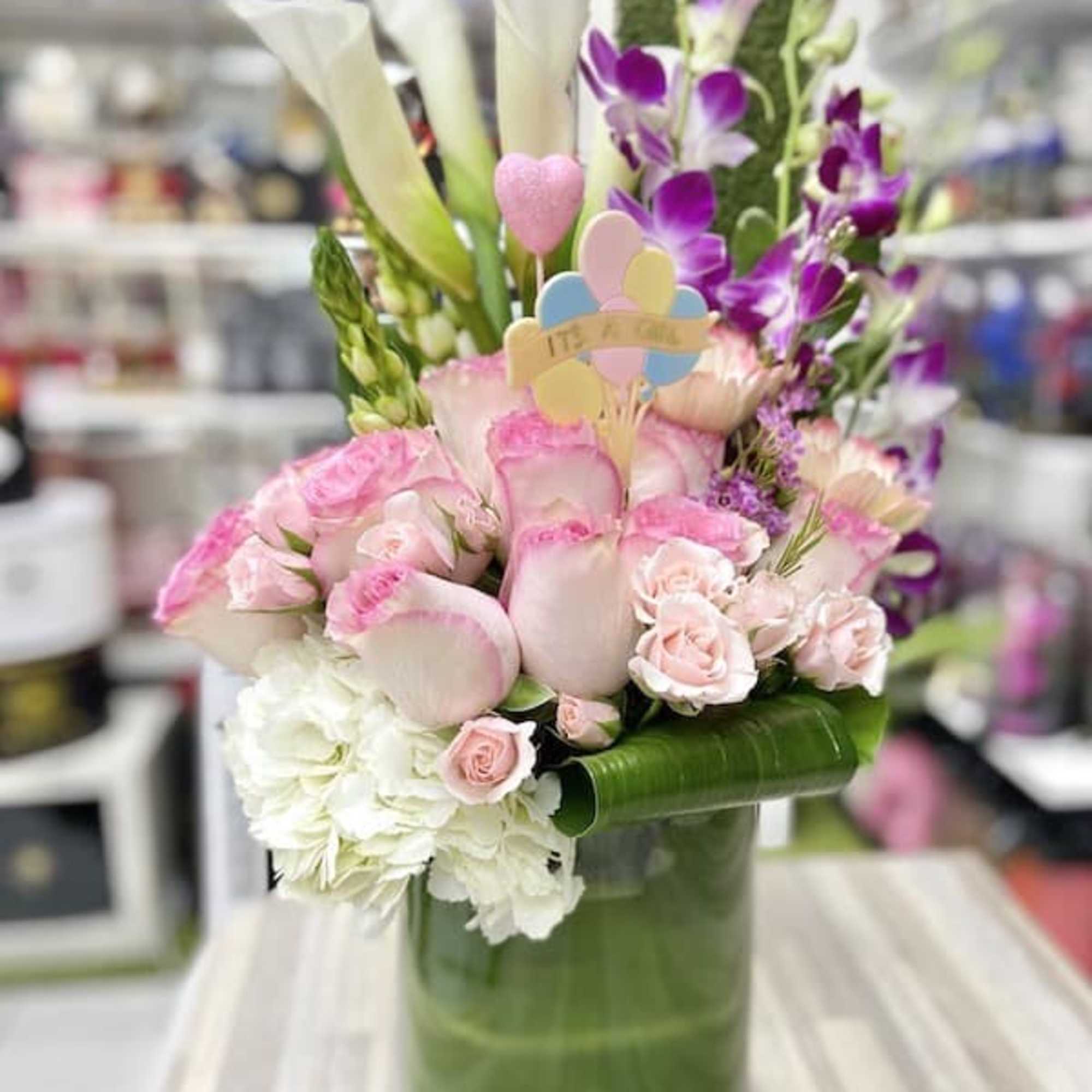 Tall arrangement of pink and white flowers with calla lilies in a green glass vase and an 'It's a Girl' pick
