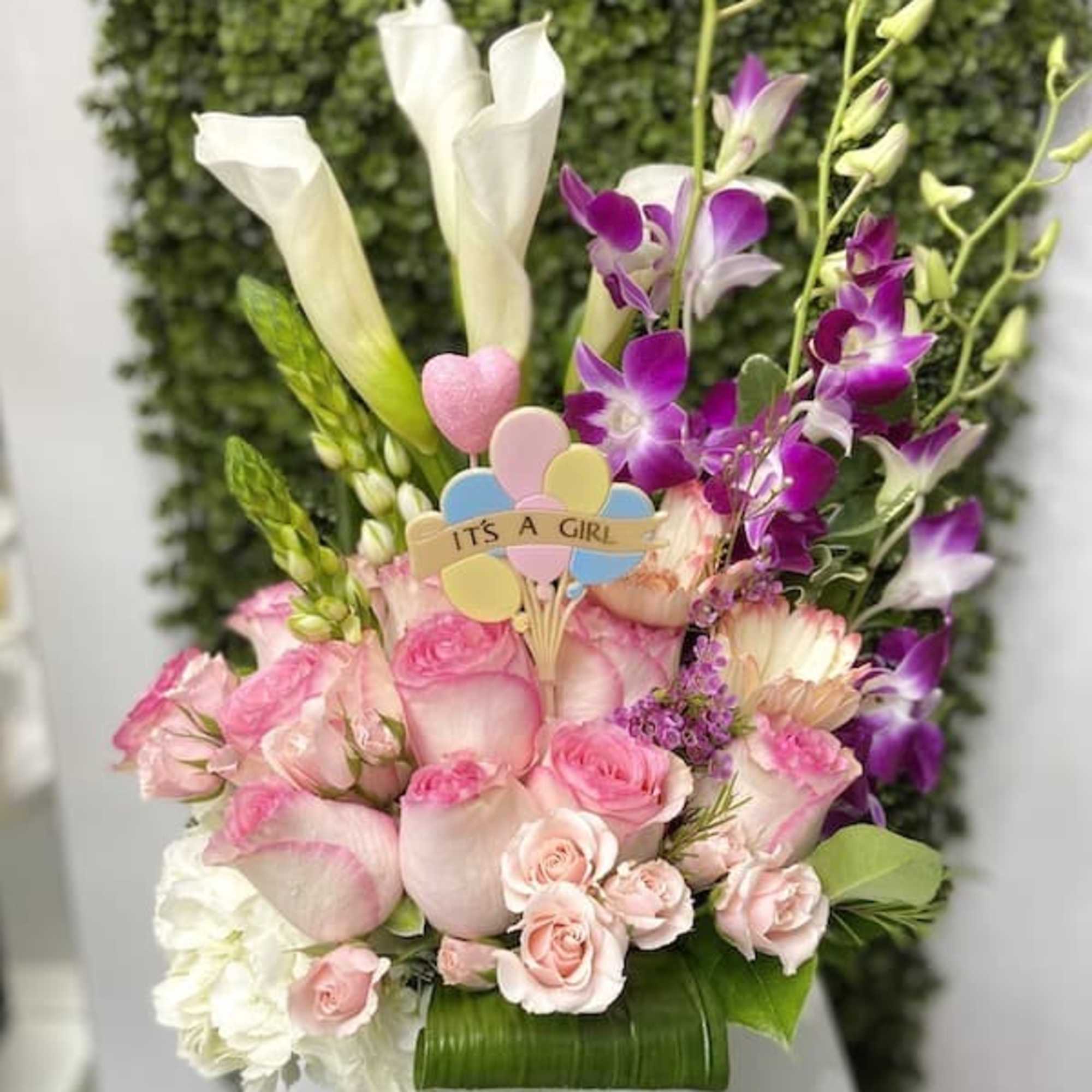 Pink and white flower arrangement with calla lilies, roses, orchids, and an 'It's a Girl' pick in a glass vase.