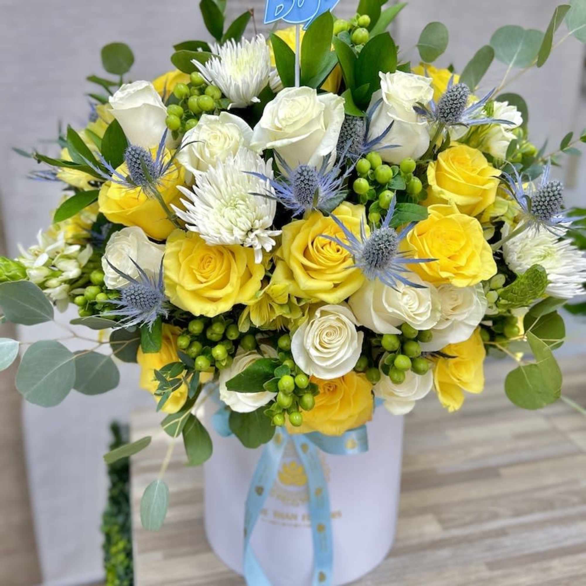 Hatbox arrangement of yellow and white roses with mums, blue thistles, and an It’s a Boy pick.