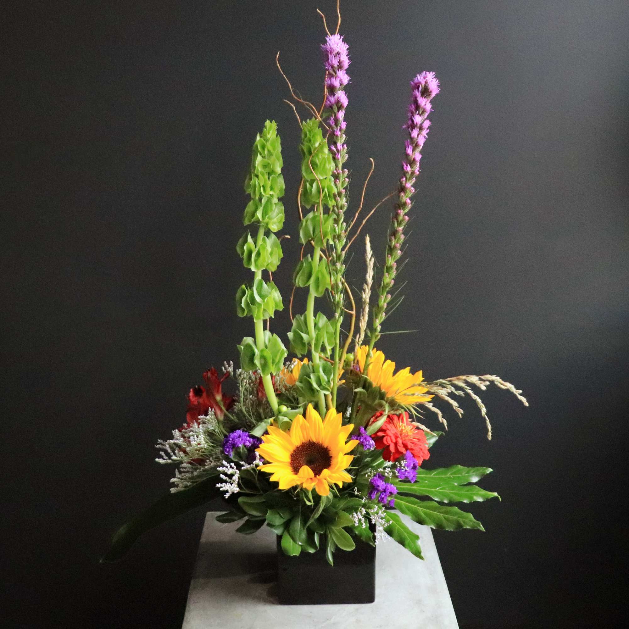 Tall mixed flower arrangement with sunflowers, purple spikes, and red blooms in a black container