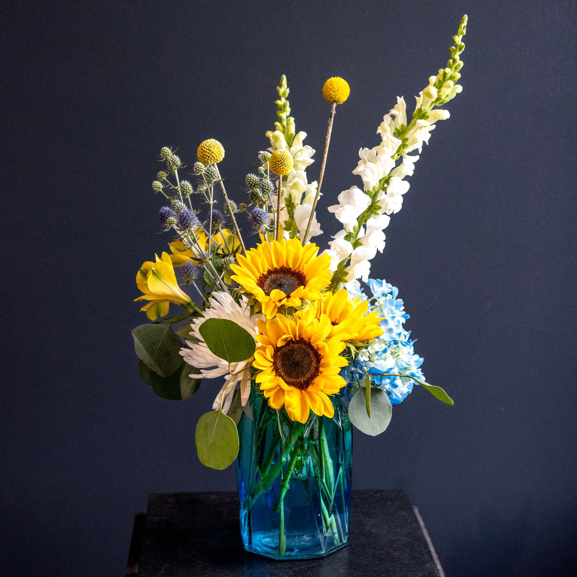 Sunflowers, white snapdragons, and blue hydrangeas in a blue glass vase
