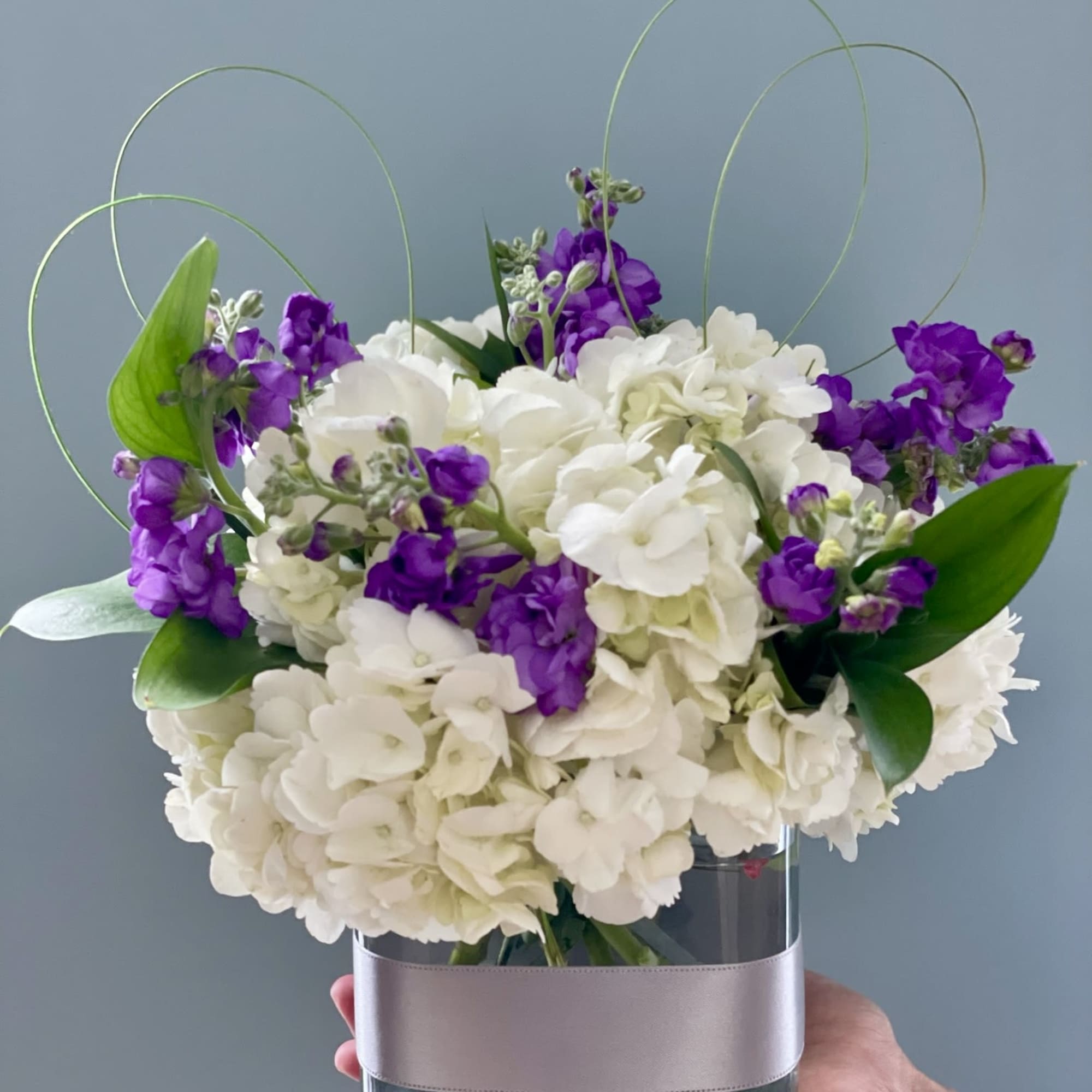 White hydrangeas and purple stock in a glass vase with looping wire accents