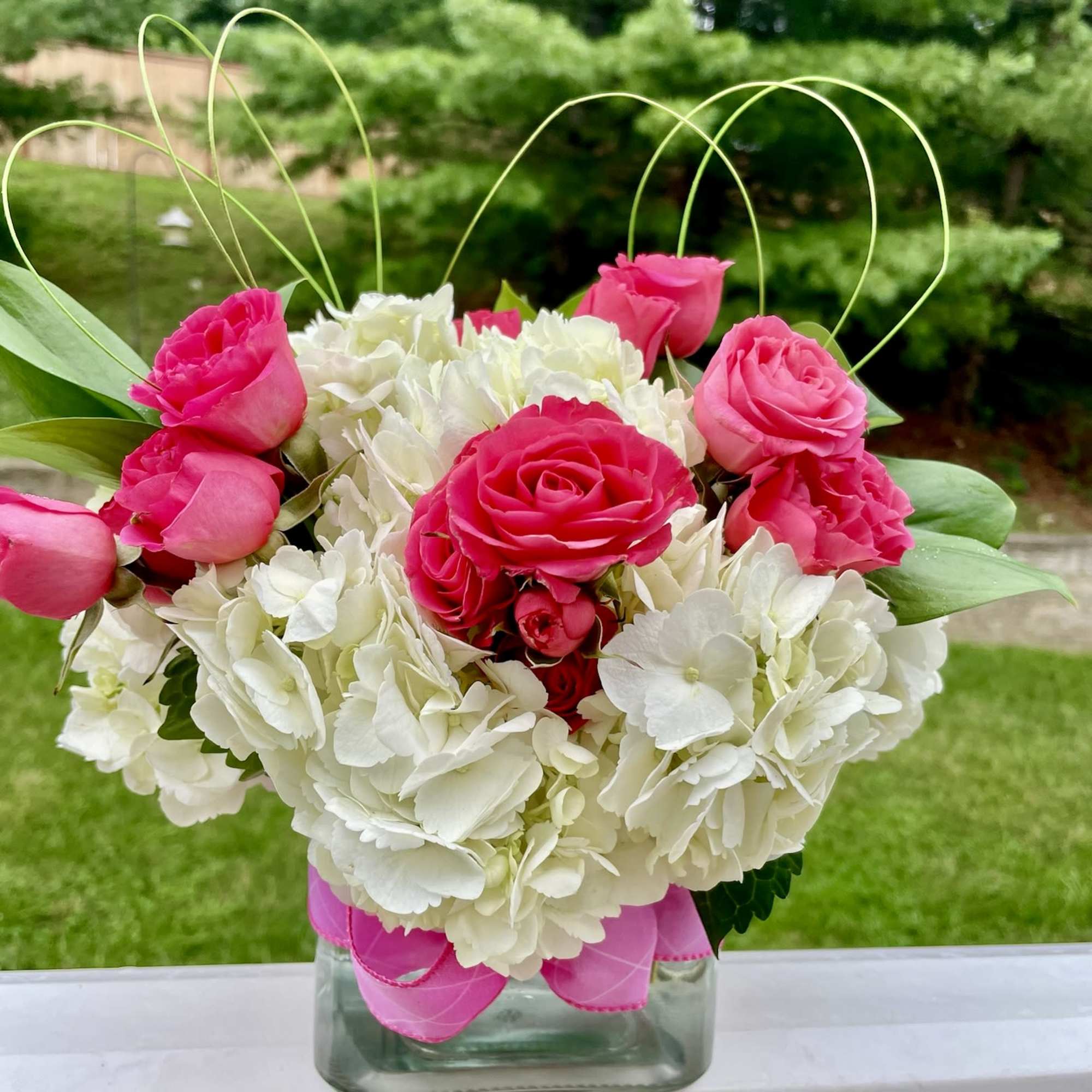 Pink roses and white hydrangeas arranged in a glass vase with a pink ribbon.