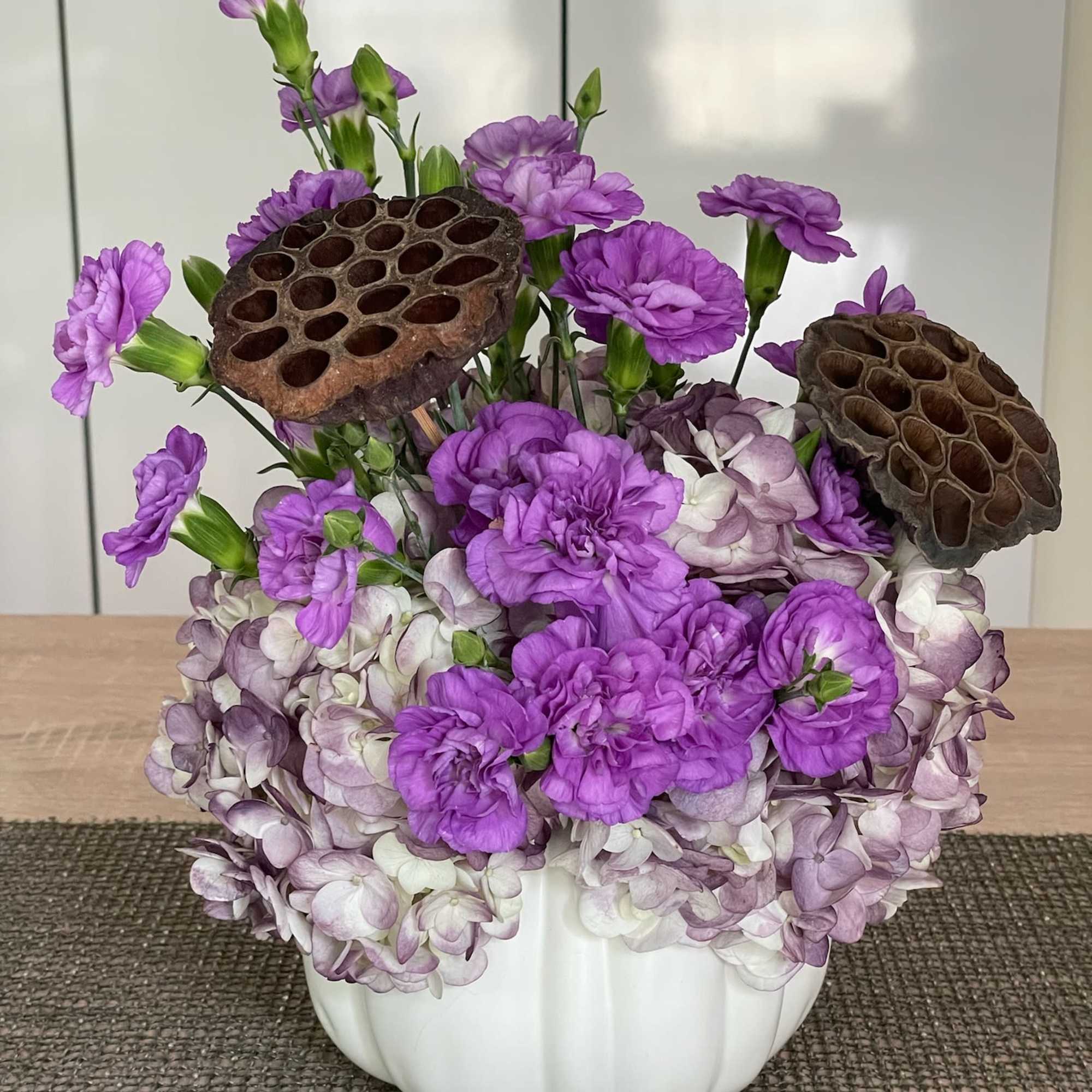 Purple carnations and hydrangeas in a white pumpkin vase with lotus pods