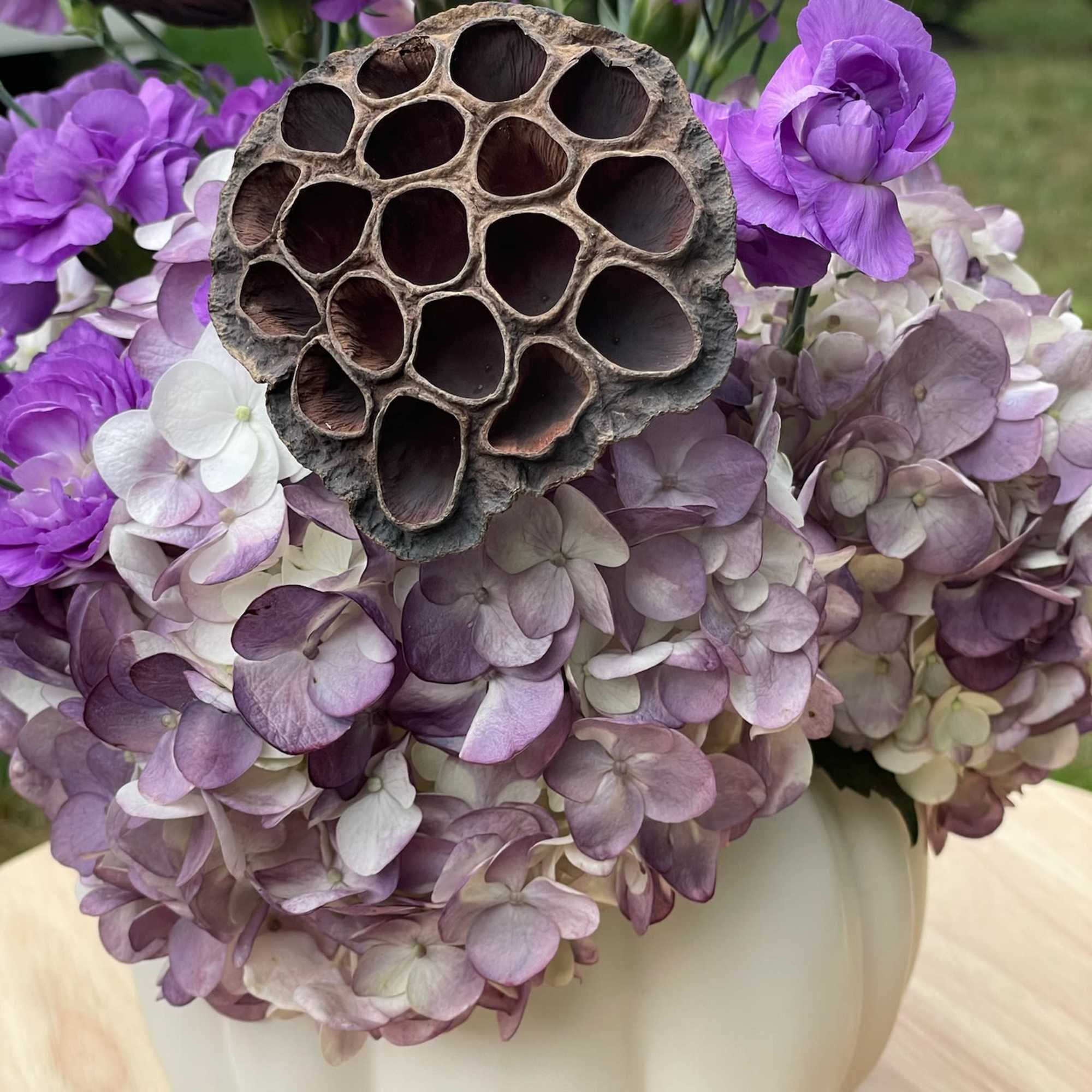 Purple flowers and hydrangeas arranged in a white pumpkin vase with a lotus pod accent